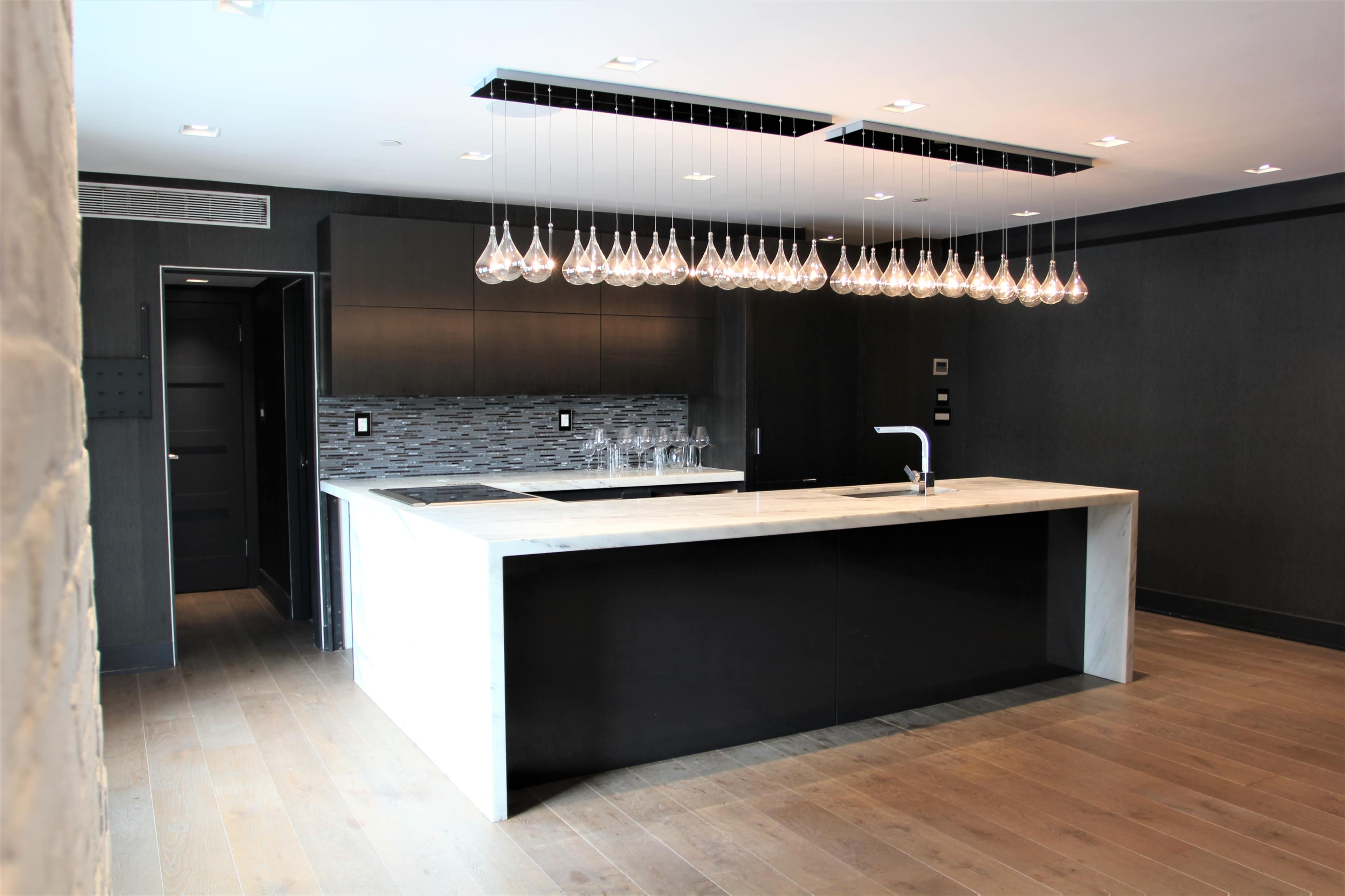 A modern kitchen features a large island with a white marble countertop and a row of pendant lights above it.