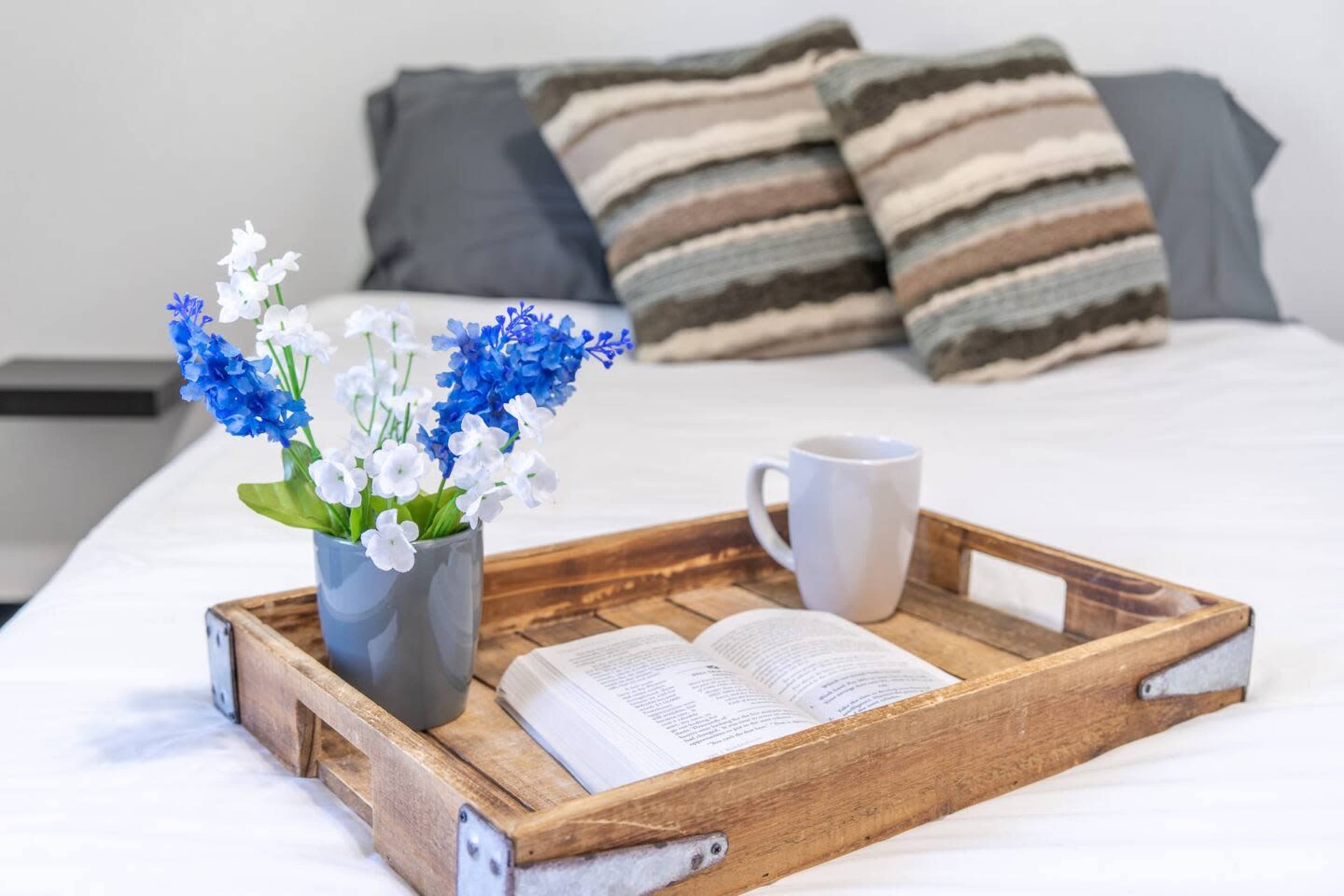 A wooden tray holds a cup and an open book next to a potted plant with blue and white flowers on a neatly made bed with patterned pillows.