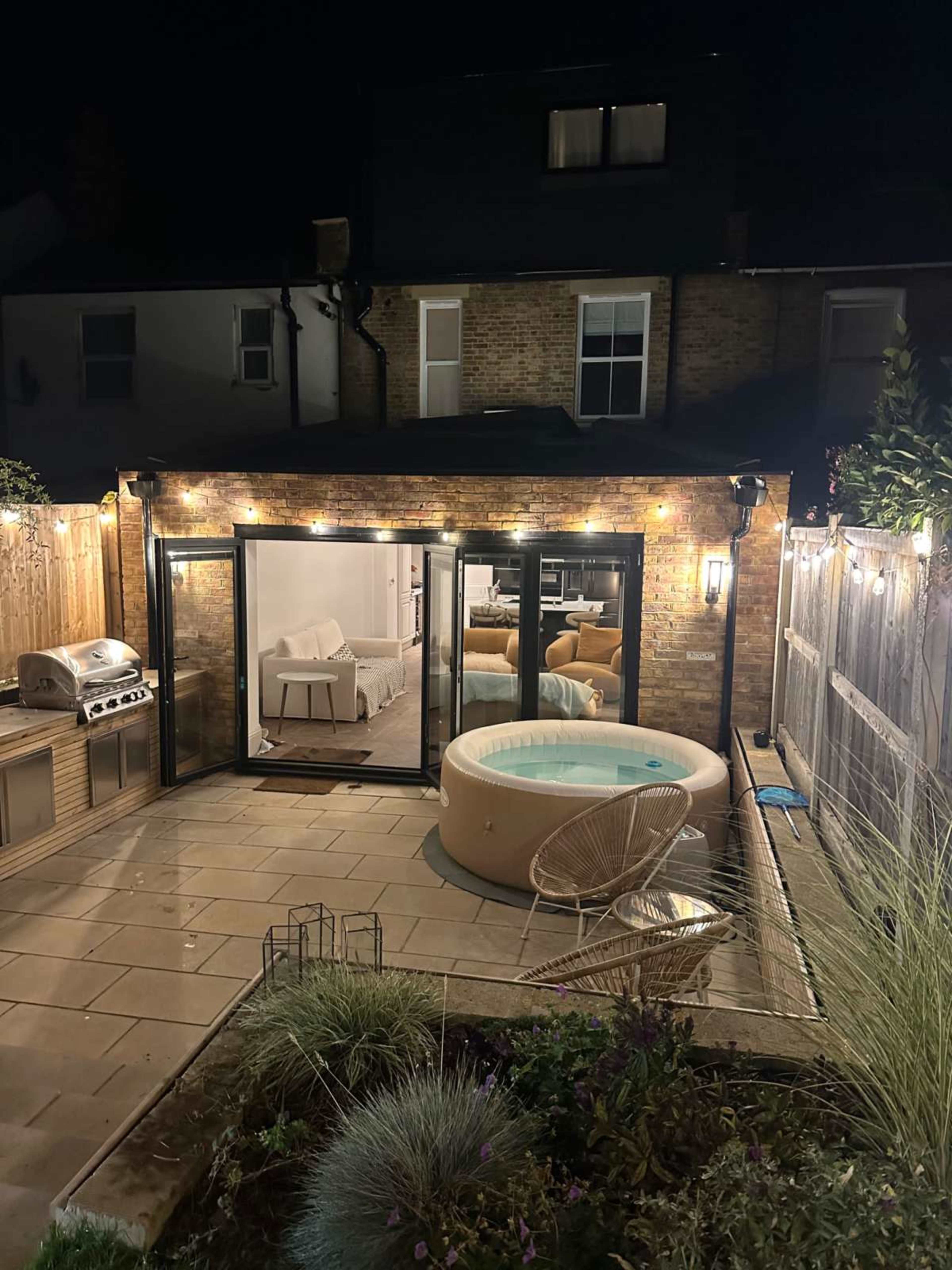 The image shows a well-lit outdoor patio with a hot tub, a barbecue grill, and an adjoining room visible through large glass doors at night.