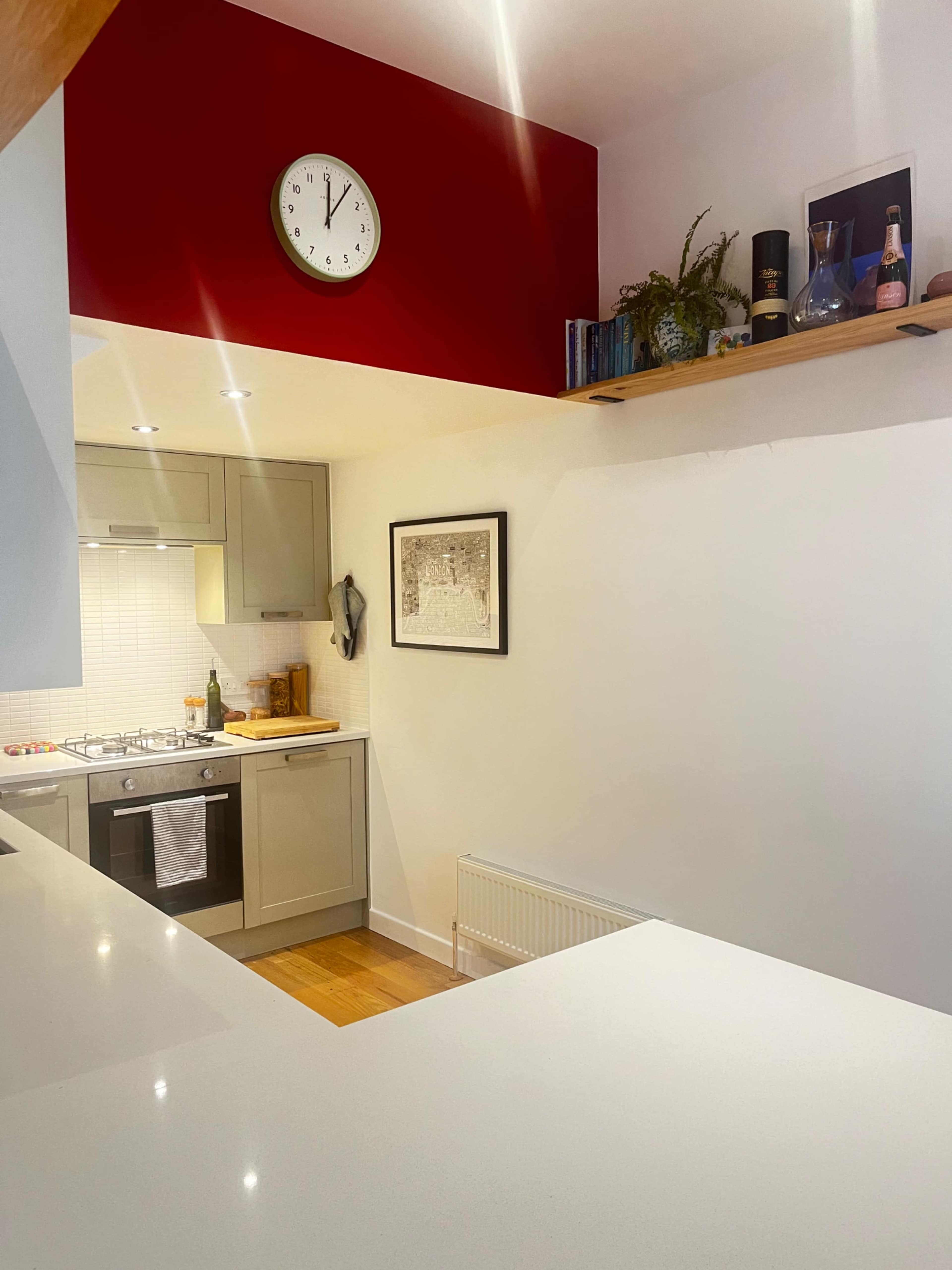 A modern kitchen with light-colored cabinetry, a clock on the red wall, and a wooden shelf displaying decorative items.