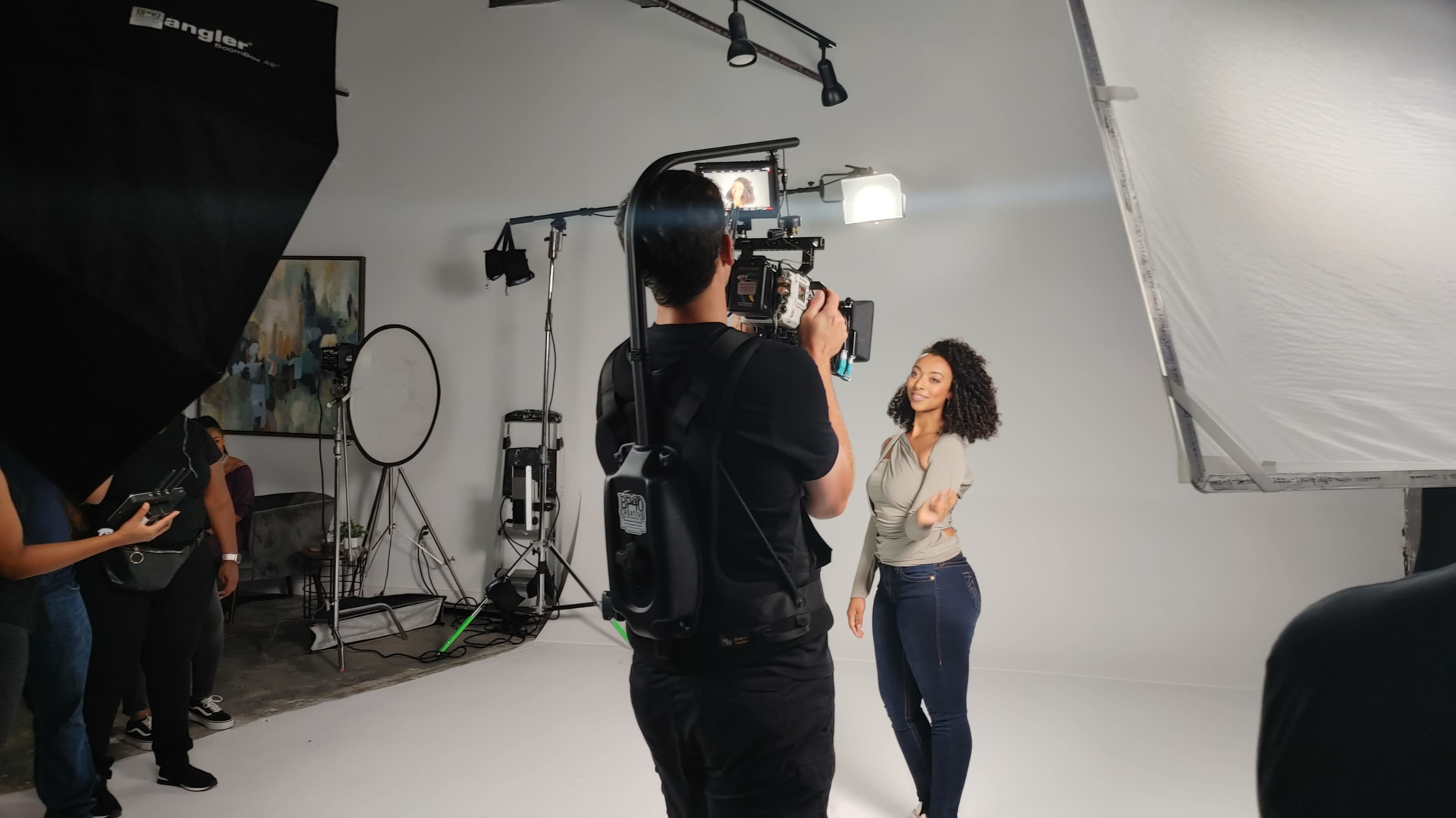 A person is filming a woman posing in a studio setting with various lighting equipment and a backdrop.
