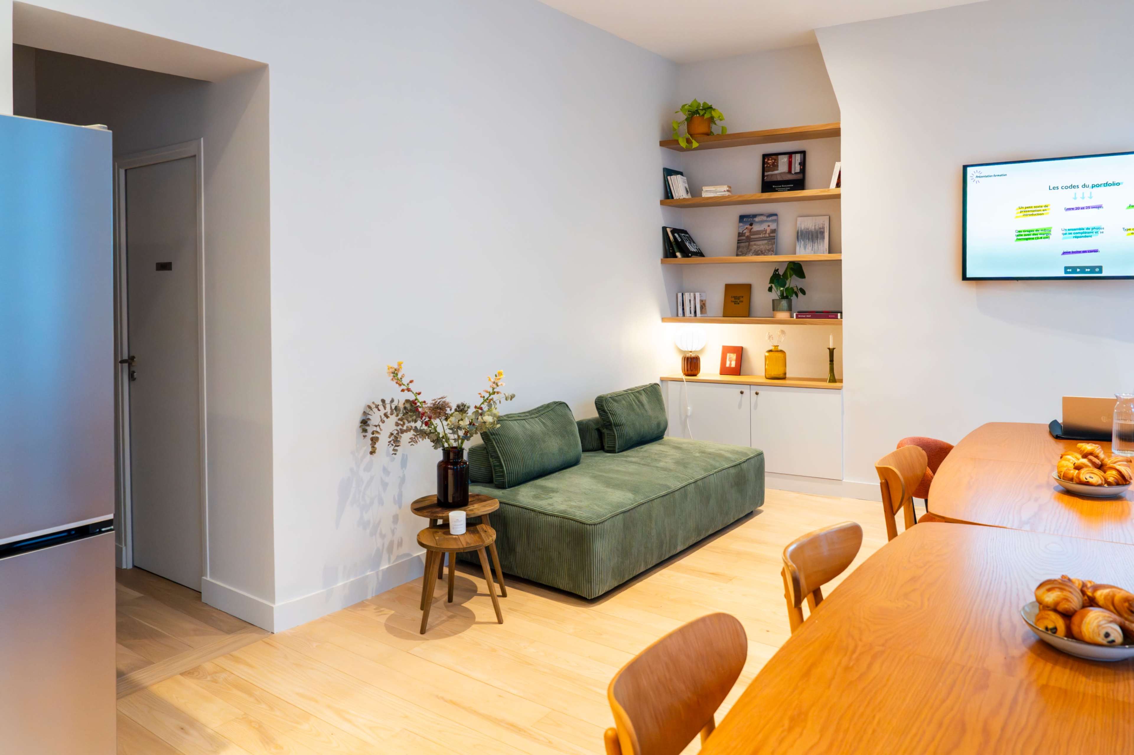 The image shows a modern interior space with a green couch, wooden dining table, and shelves displaying books and decorative items.
