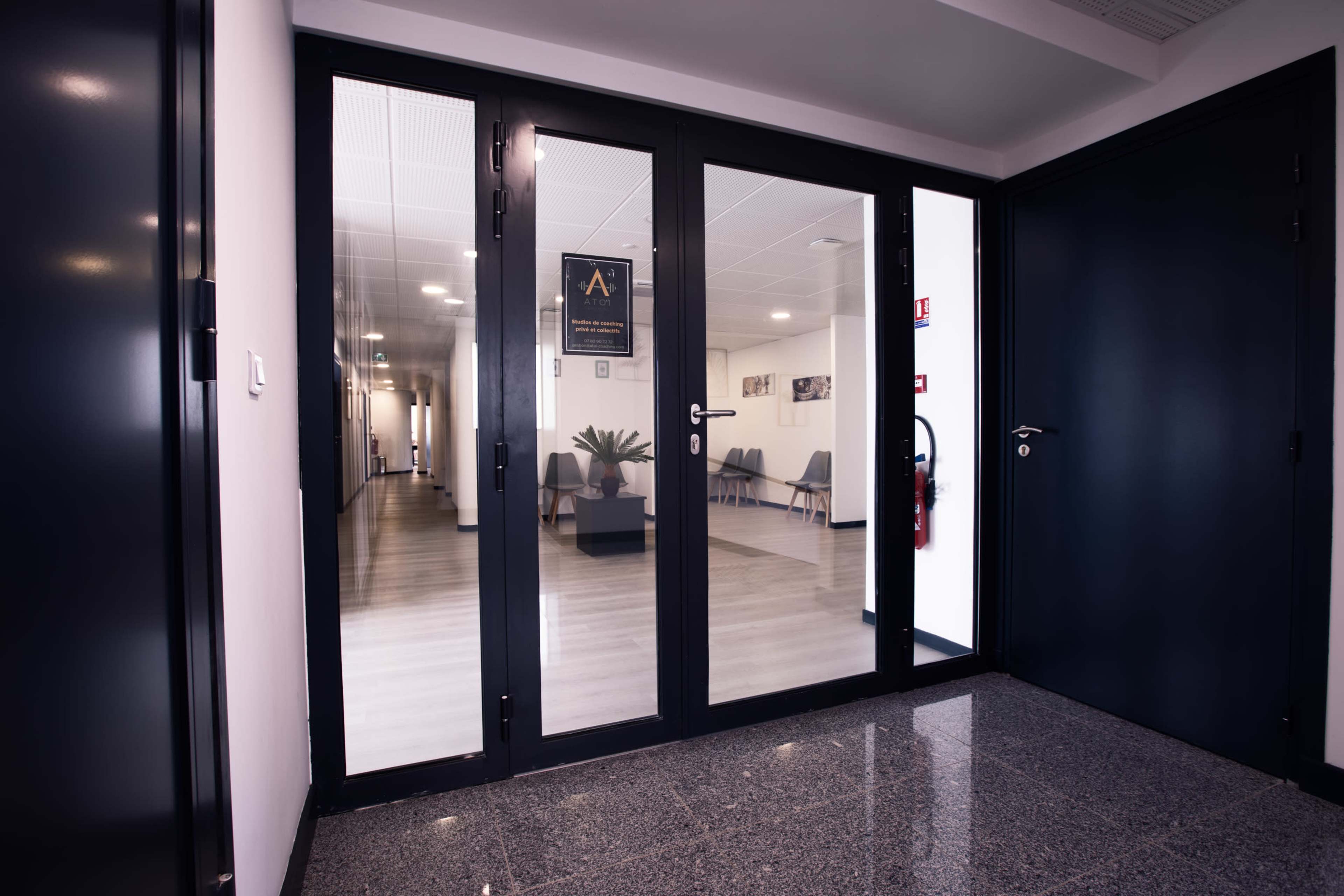A hallway with glass doors leads into a well-lit office space featuring a seating area and contemporary decor.