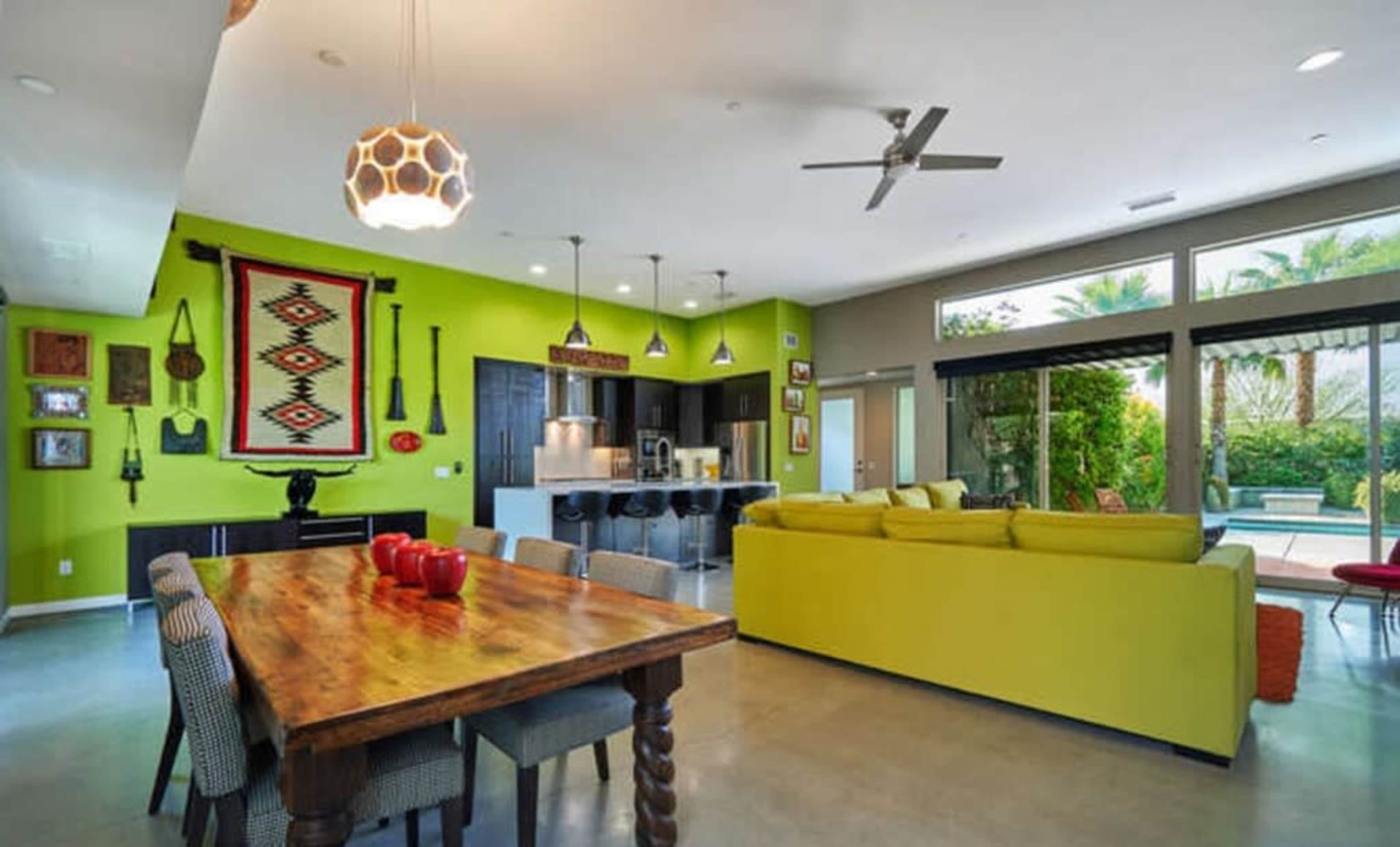 A modern living space with a green accent wall, a wooden dining table, a bright yellow sofa, and large windows overlooking a patio and pool area.