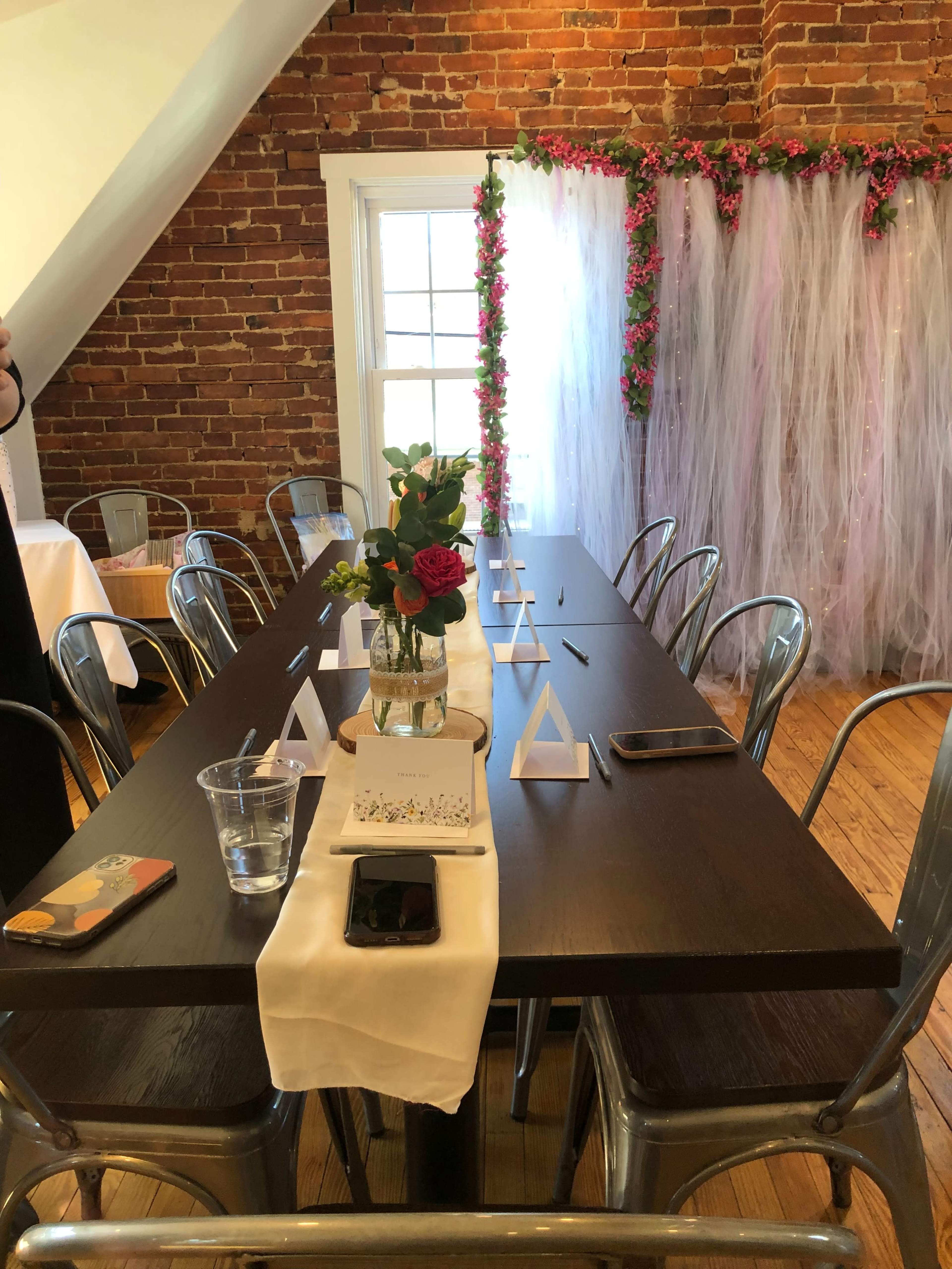 A long wooden table is set with clear chairs, a vase of roses, and decorative triangles, against a backdrop of brick walls and a floral arrangement.