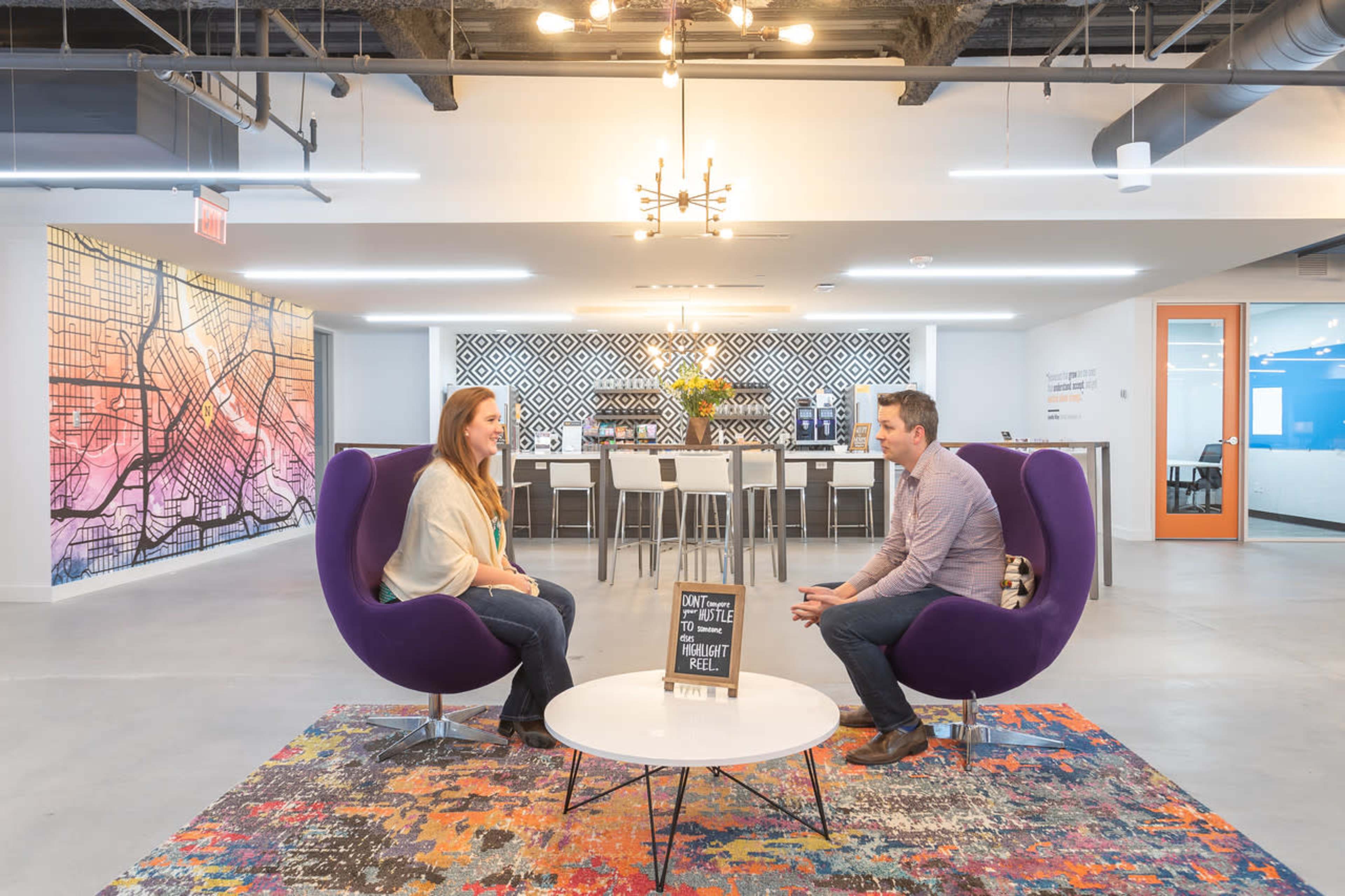 Two people sit in purple chairs facing each other in a modern office space decorated with a patterned wall and a colorful rug.