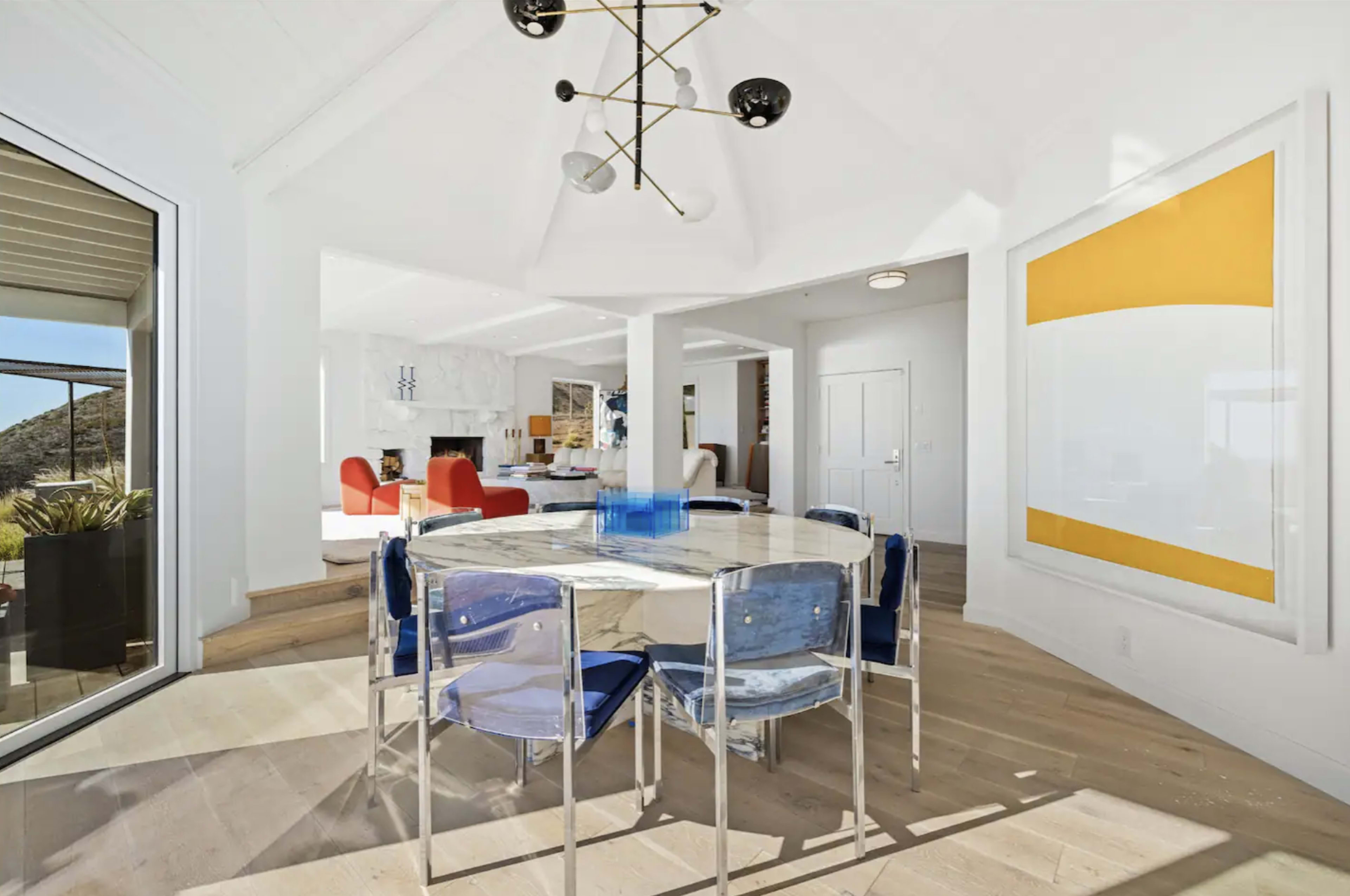 A modern dining area features a glass table surrounded by blue and clear chairs, with a view of a bright living space beyond.
