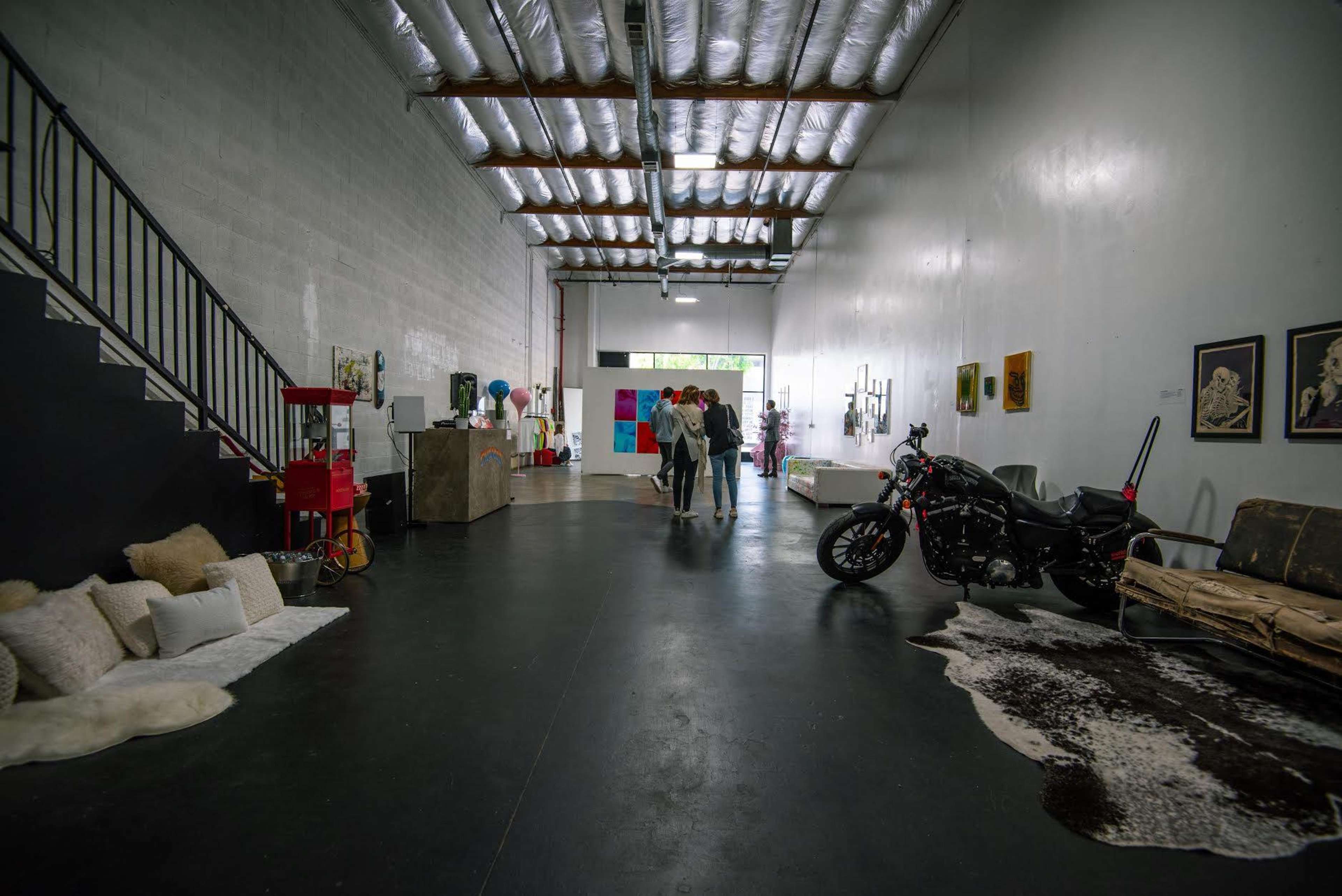 A spacious interior of an art gallery features a motorcycle, various artworks on the walls, and a couple walking through the center of the room.