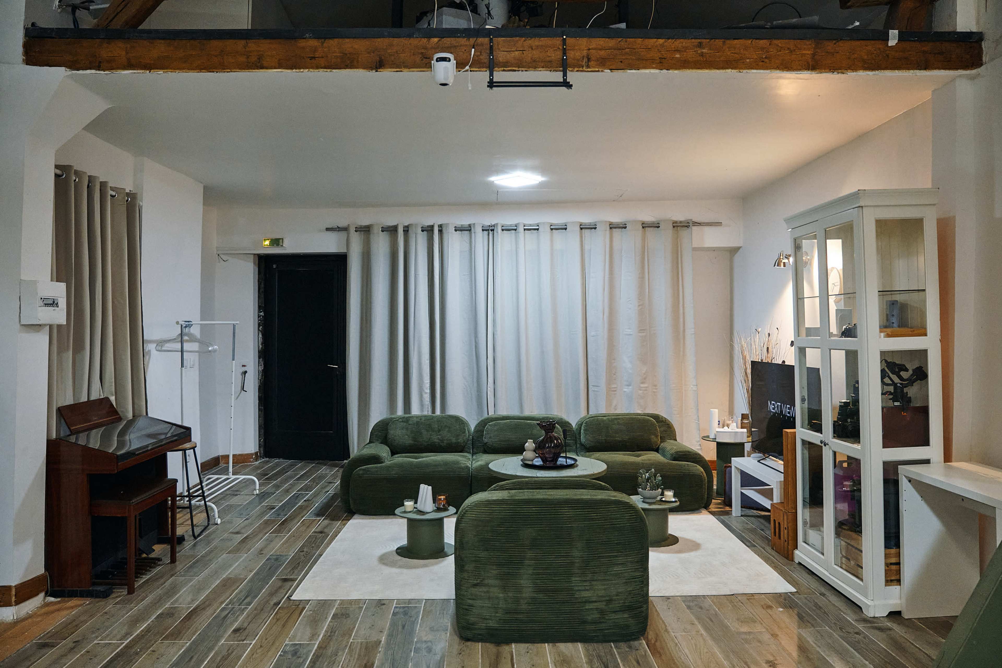 The image shows a modern, open living space featuring a green seating arrangement, a small round table, and wooden flooring, illuminated by soft lighting.