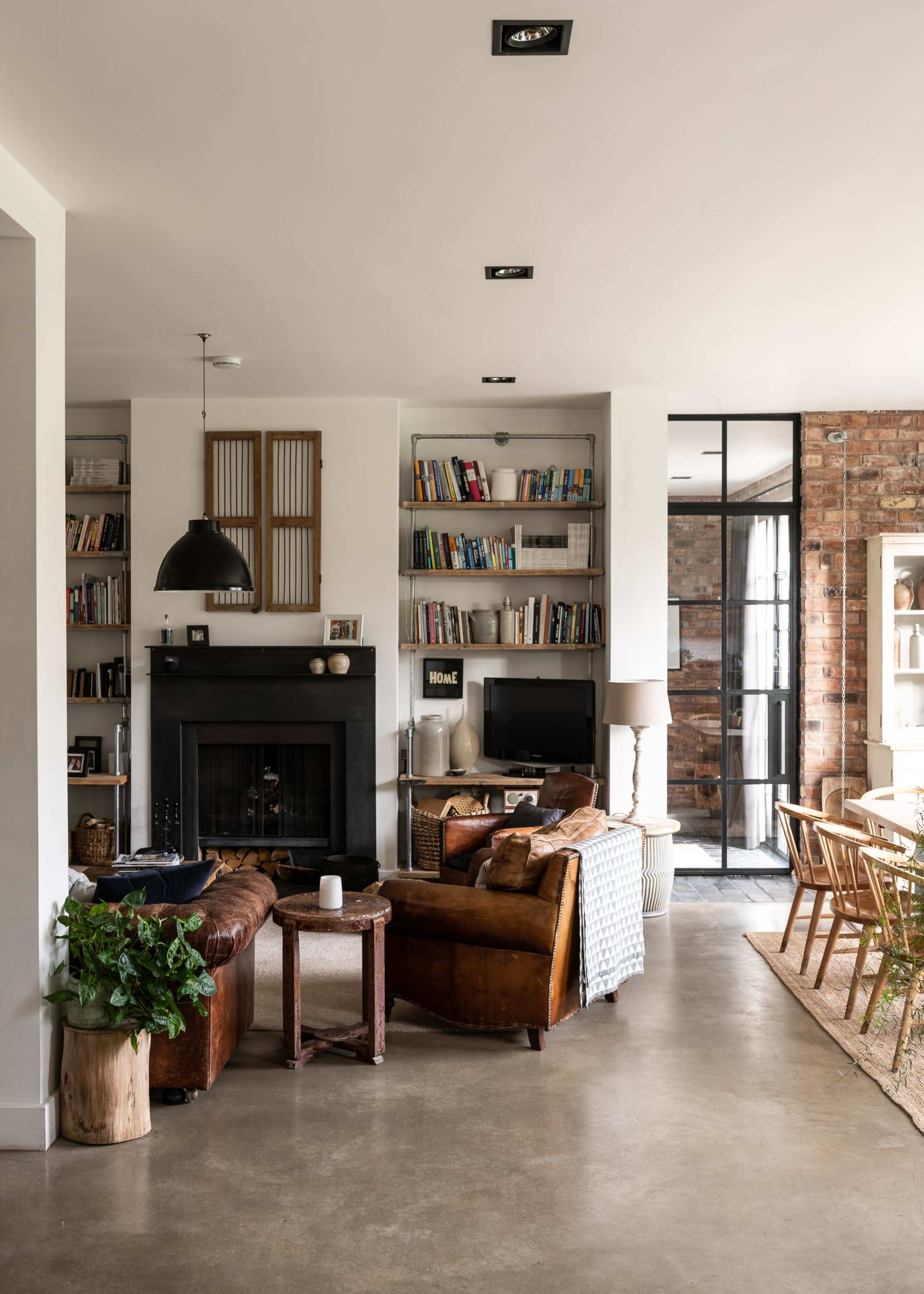 A modern living room featuring a fireplace, bookshelves filled with books, leather furniture, and a dining area with large windows and a wooden table.