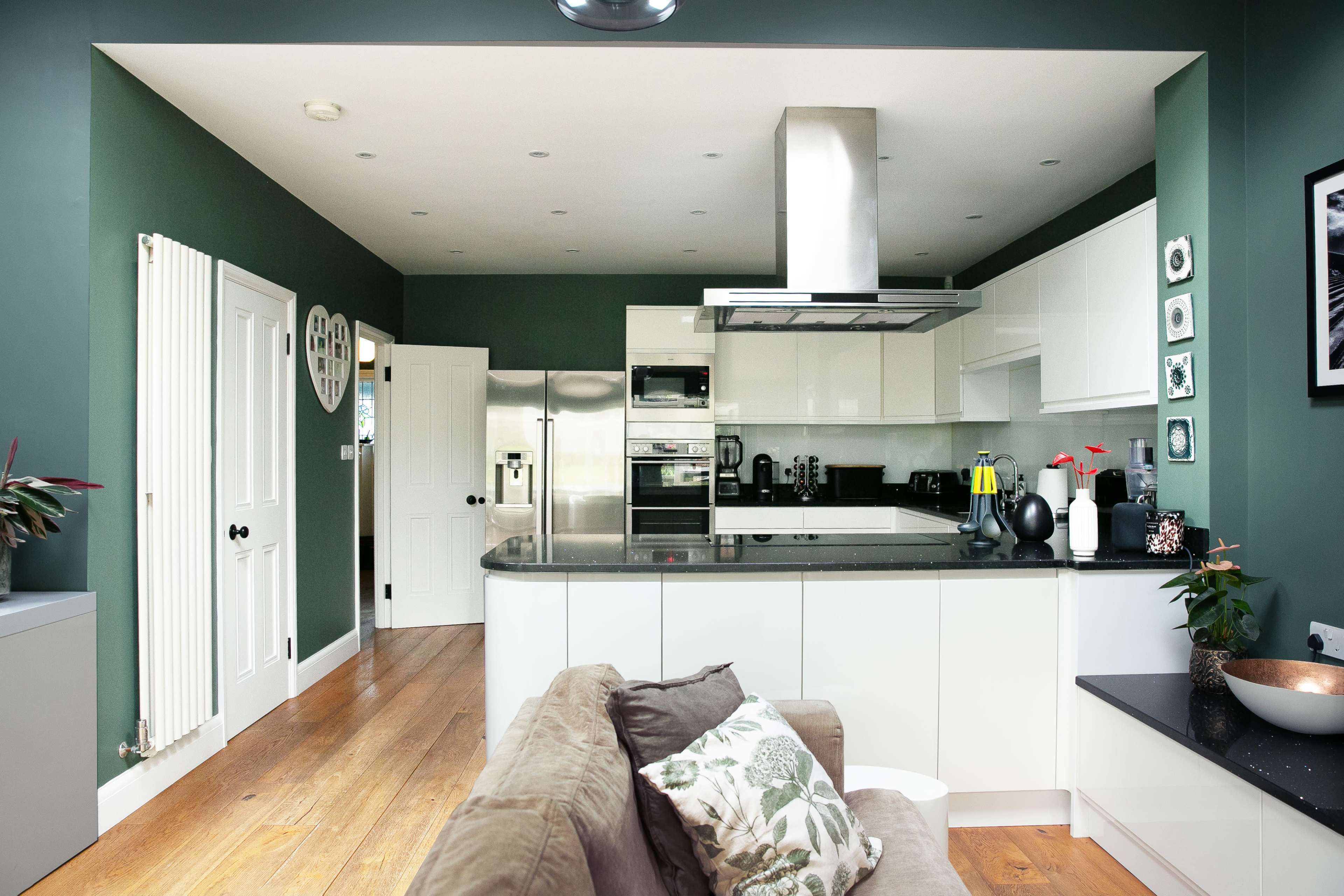 The image shows a modern kitchen with green walls, white cabinetry, and hardwood flooring, featuring a central island and stainless steel appliances.