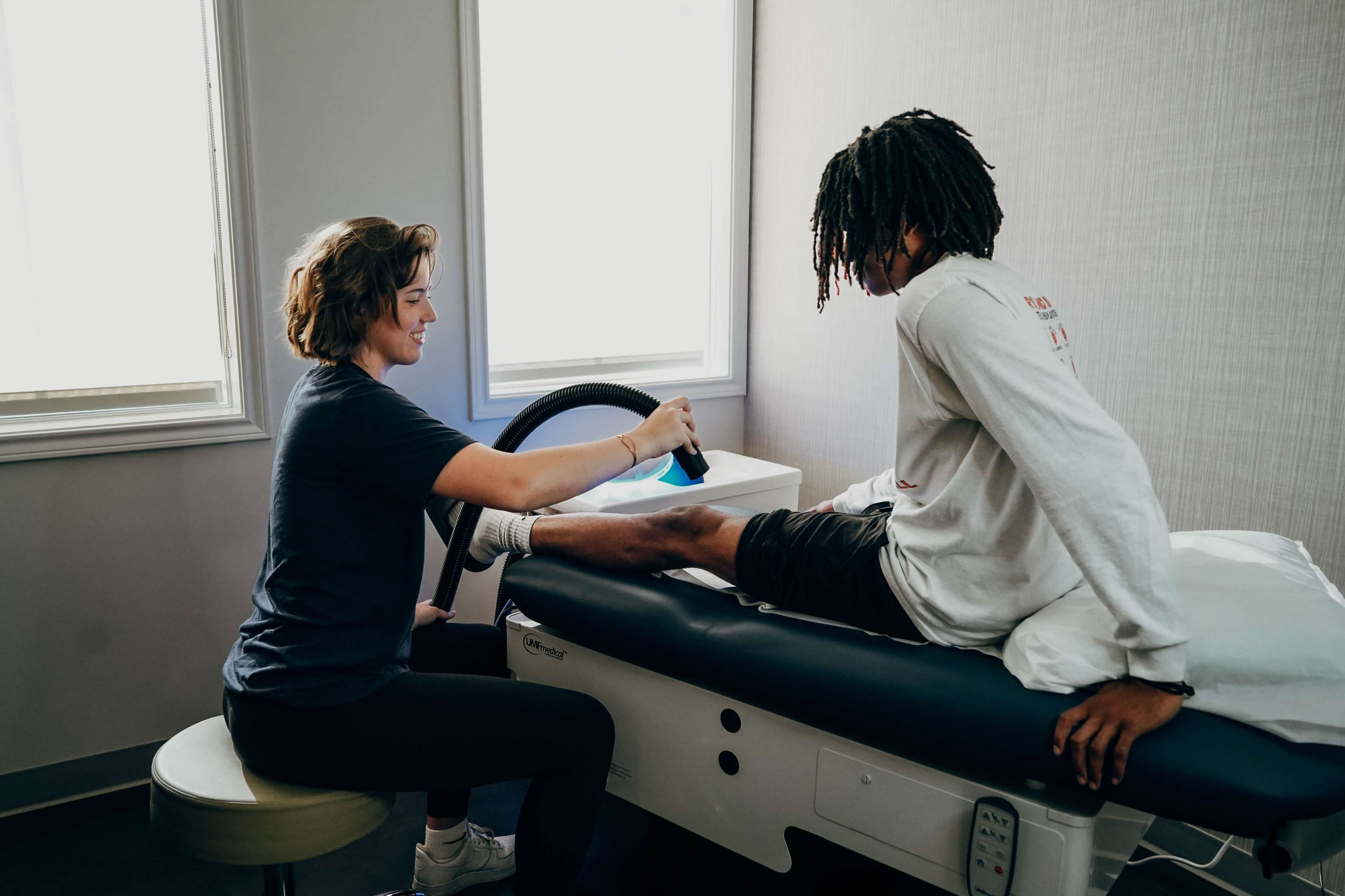 A therapist uses a device on a patient's leg during a treatment session in a clinical setting.
