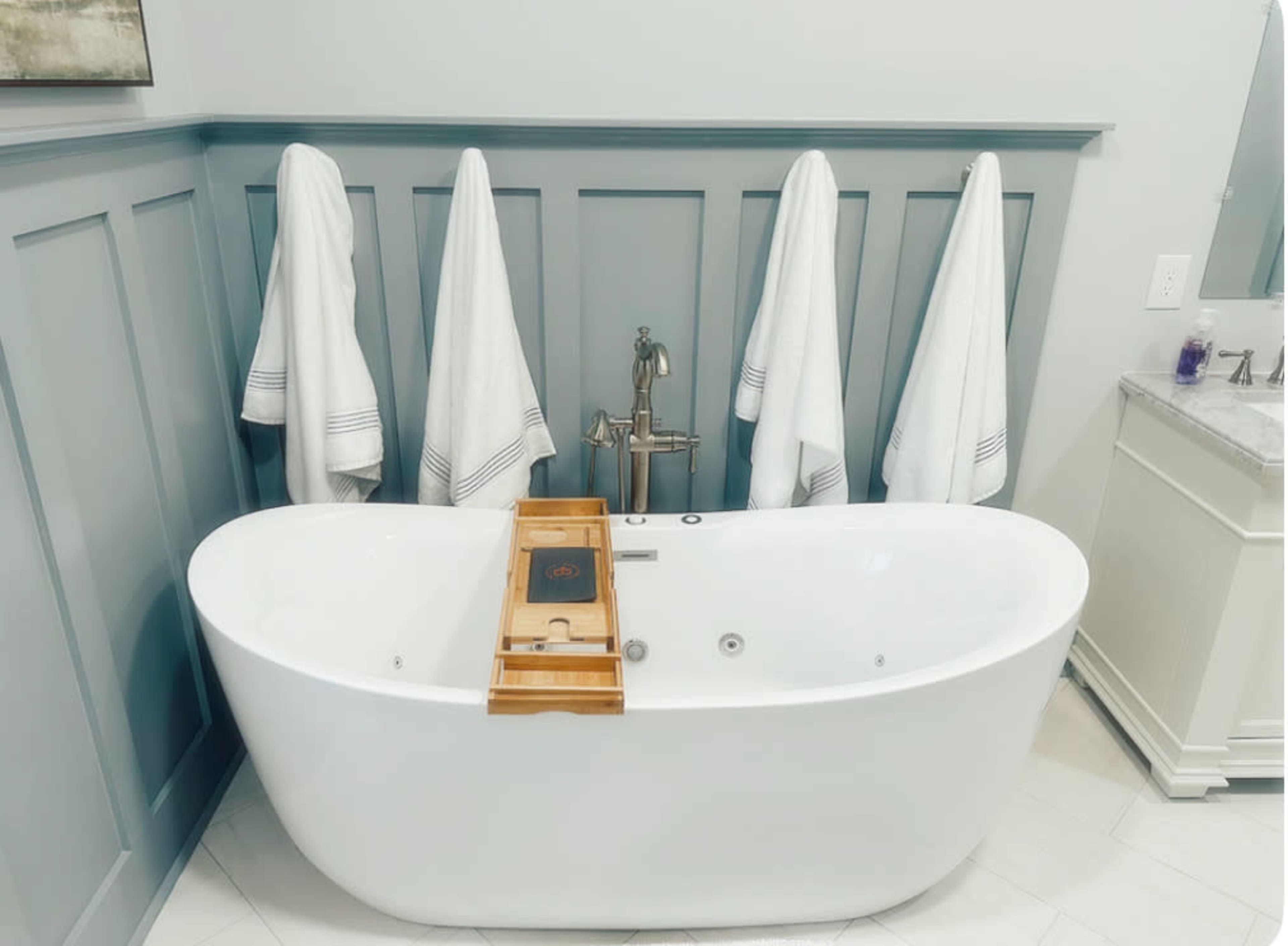A freestanding white bathtub is centered in a bathroom with gray paneled walls, flanked by four hanging white towels and a wooden bath caddy.