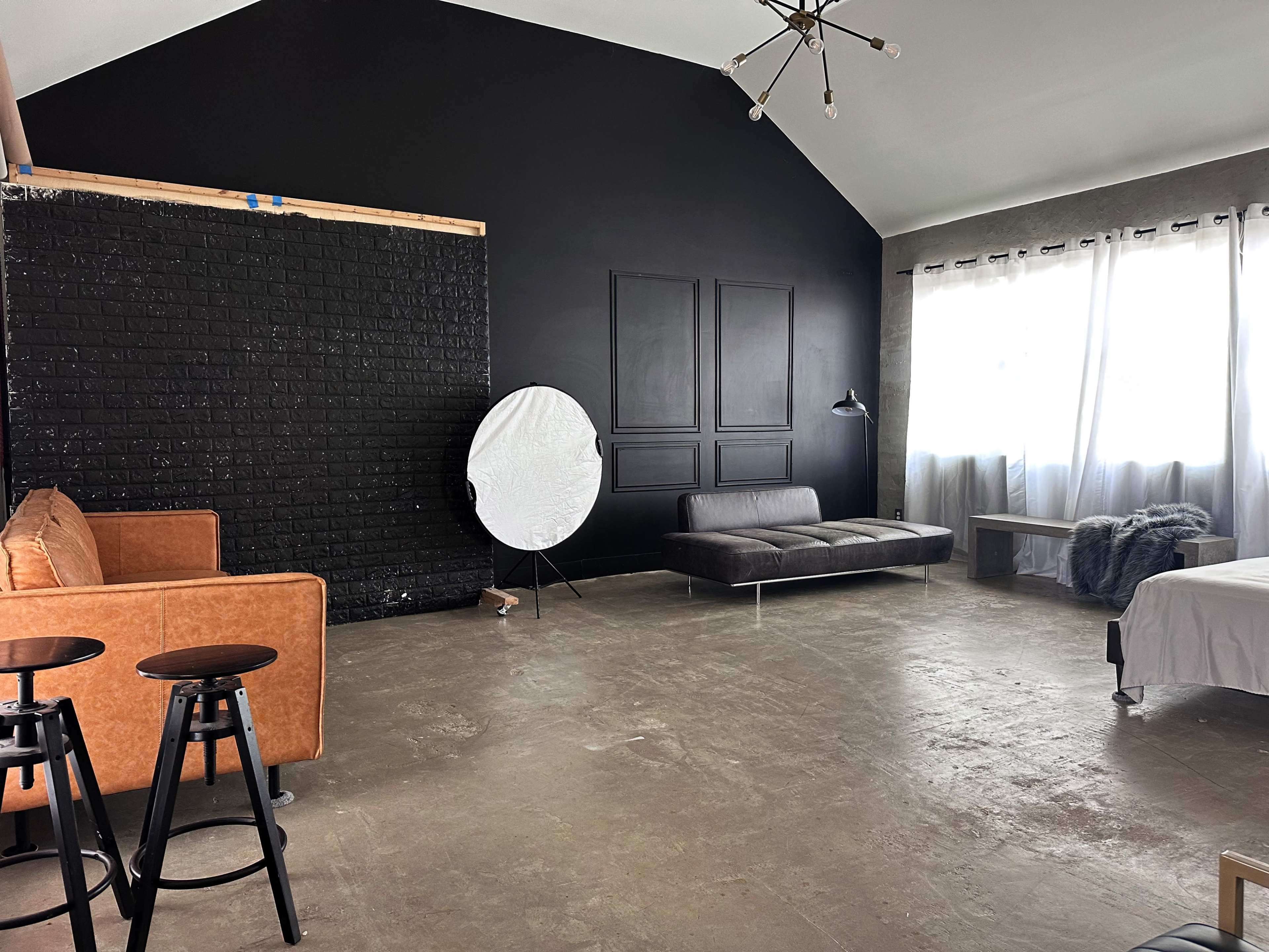 A spacious room with a black accent wall, a brown sofa, two black stools, a round reflector, and a bed near a large window.