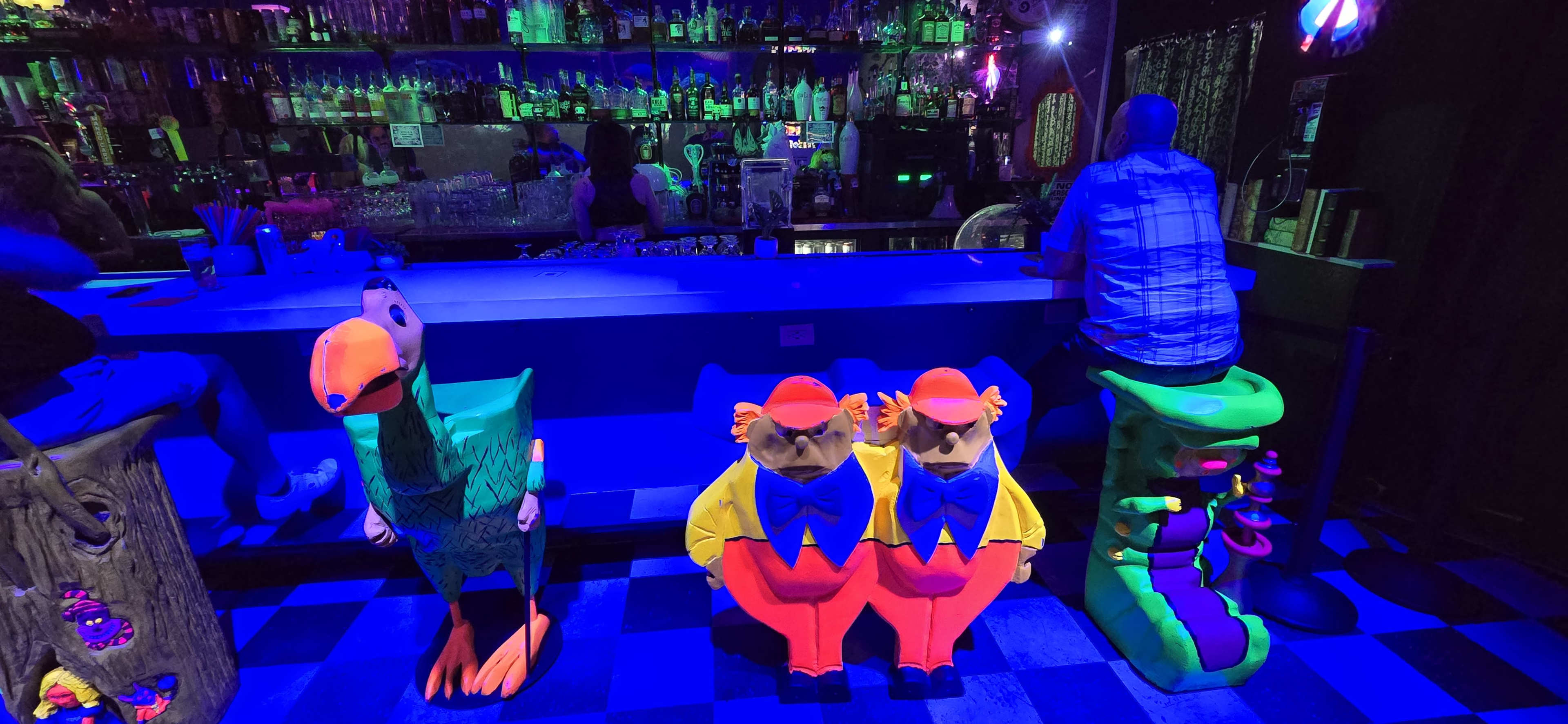 Colorful, cartoon-like characters sitting on a bar counter in front of a dimly lit bar with various bottles in the background.