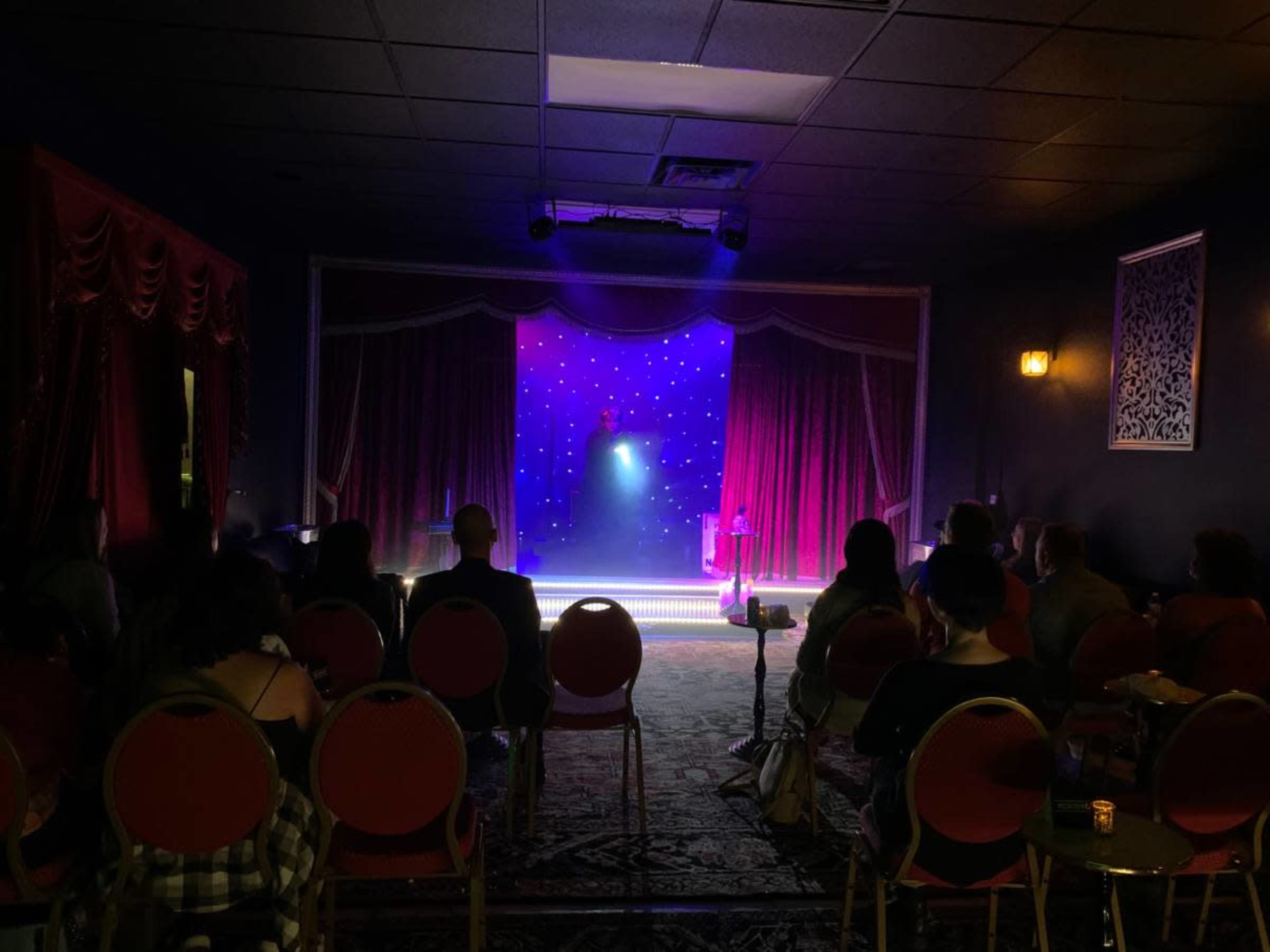 A dimly lit performance space with red curtains, a small stage illuminated by blue and purple lighting, and an audience seated in rows of red chairs.