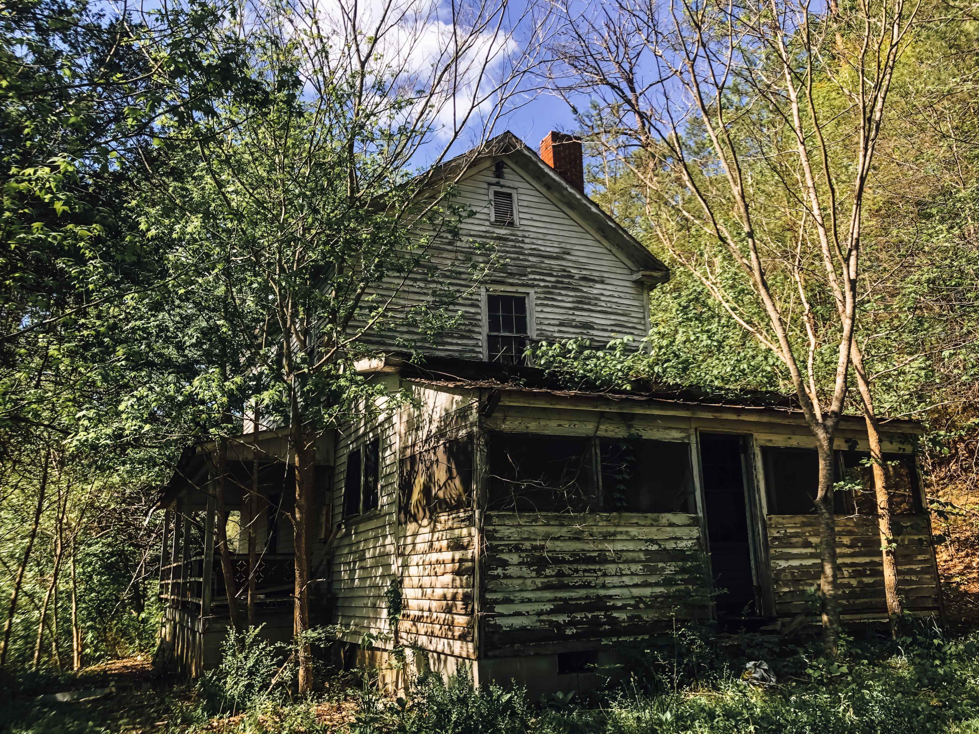 An abandoned, weathered house with peeling paint is surrounded by overgrown trees and vegetation.