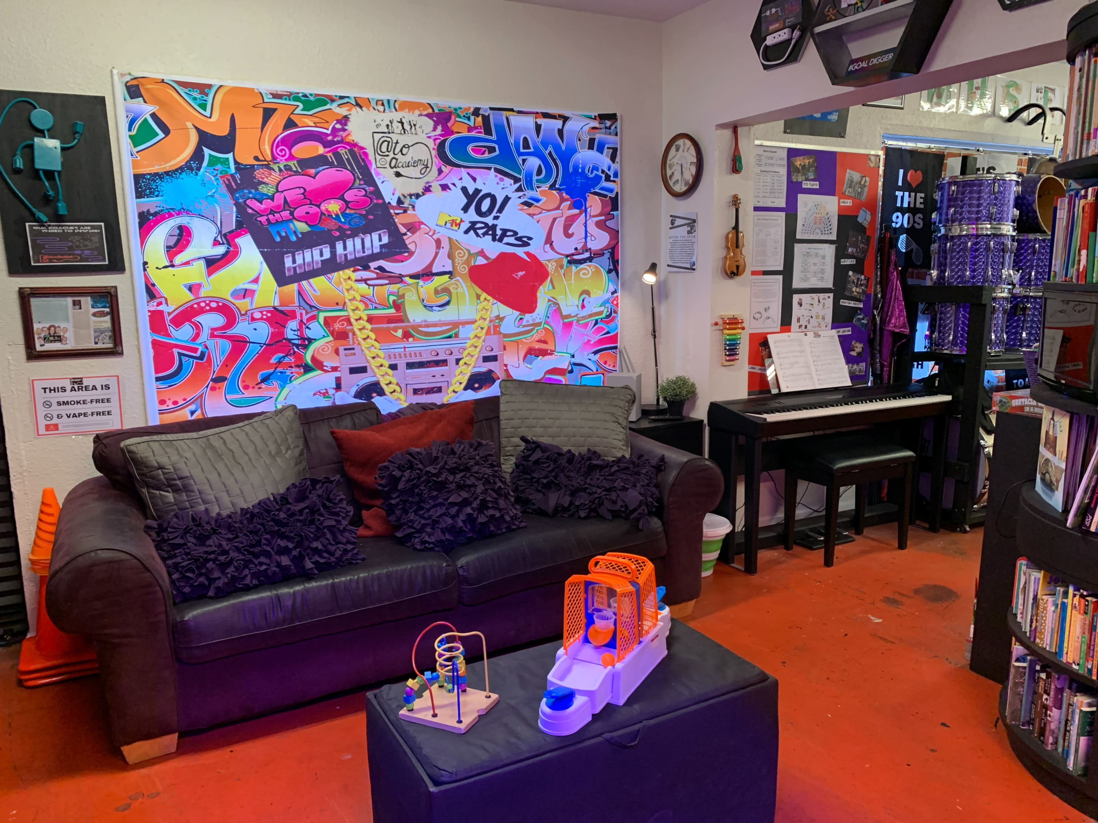 A cozy room with a dark couch adorned with pillows, a colorful graffiti mural on the wall, a small piano, and a children's toy on an ottoman.
