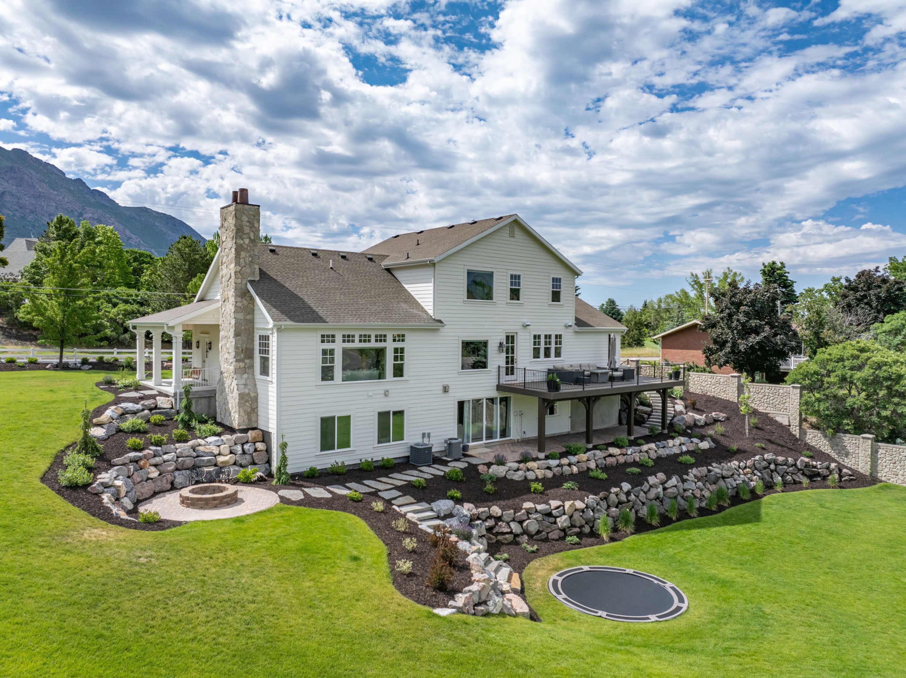 A large two-story white house with a stone chimney is surrounded by landscaped gardens and a lawn, featuring a circular trampoline in the yard.