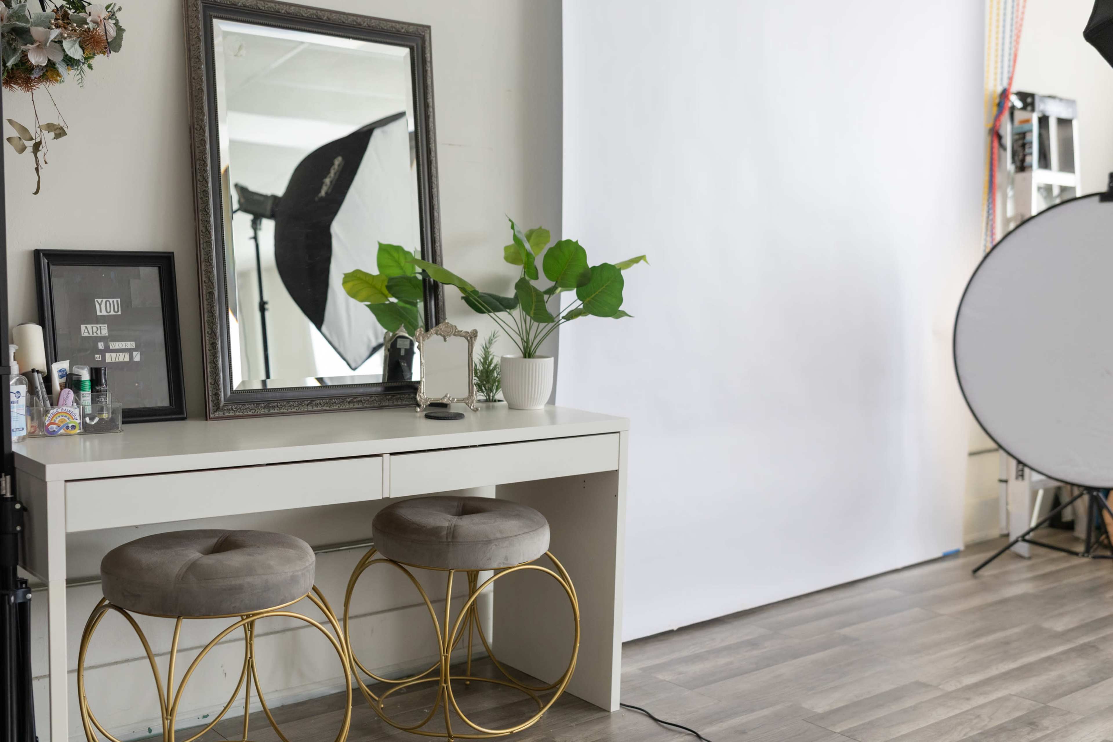 The image shows a well-lit room featuring a white backdrop with a decorative mirror and two round stools in front of a vanity table, alongside a green plant and photography equipment in the corner.