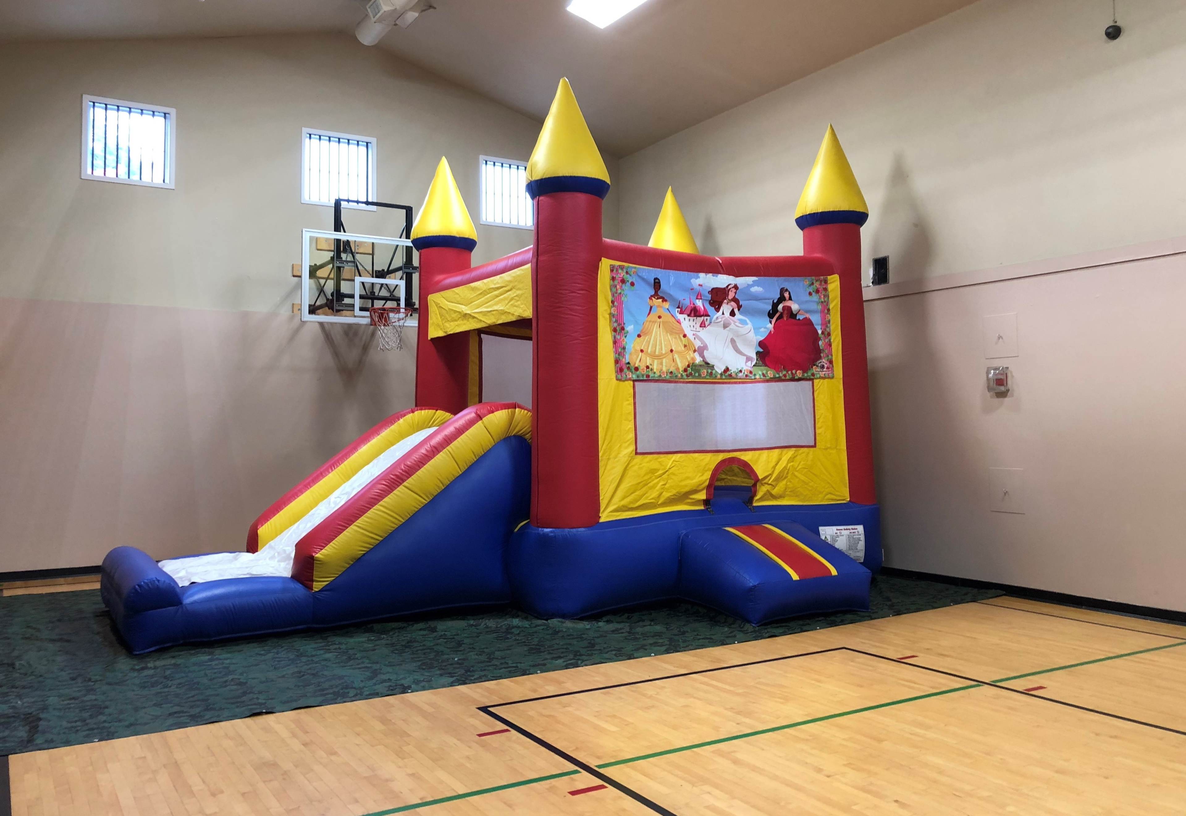 The image shows a large inflatable bounce house with a slide, set up in a gymnasium.