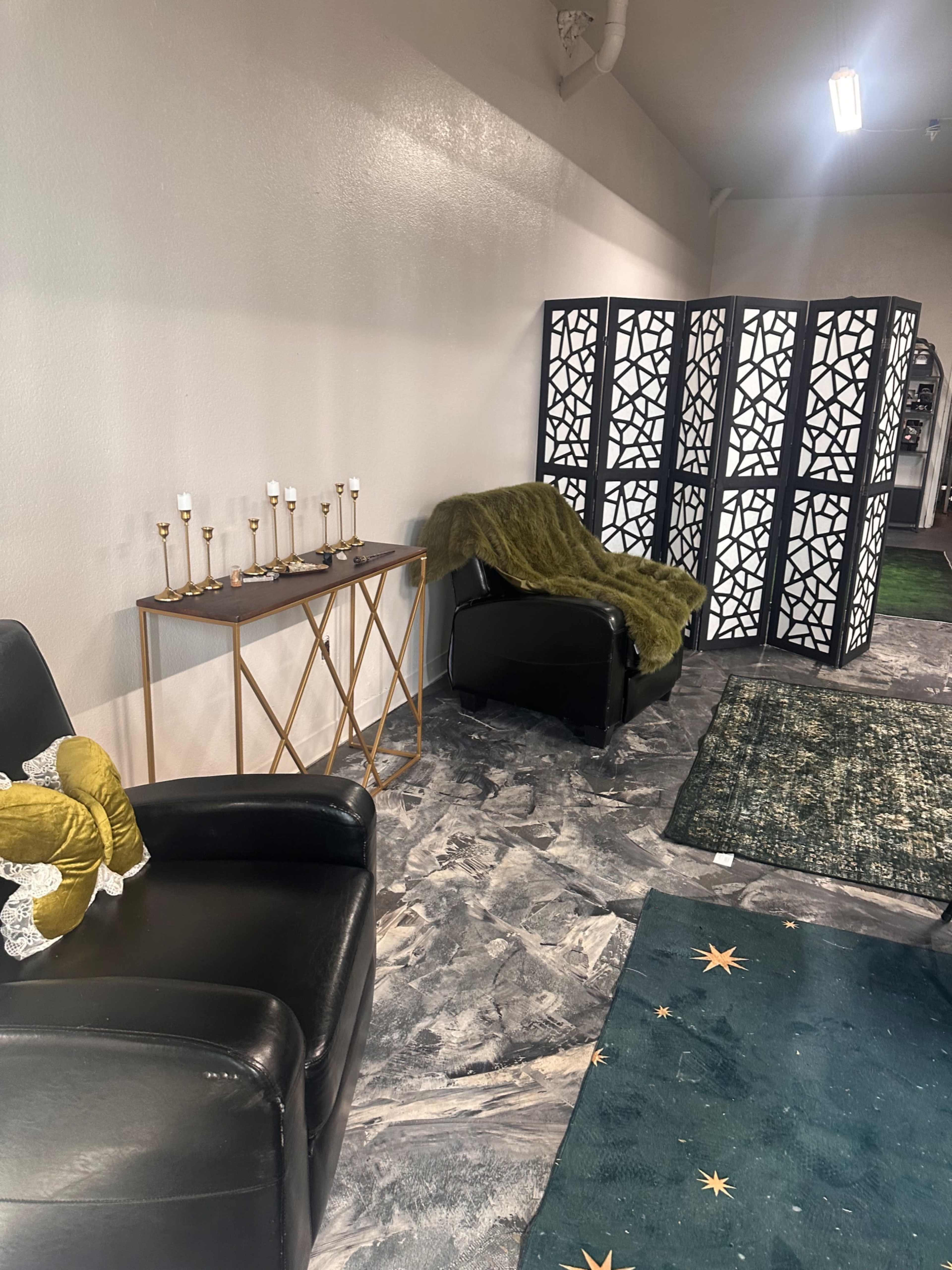 The image shows a cozy seating area with two black chairs, a green throw blanket, a decorative console table with candles, and a patterned folding screen in the background.