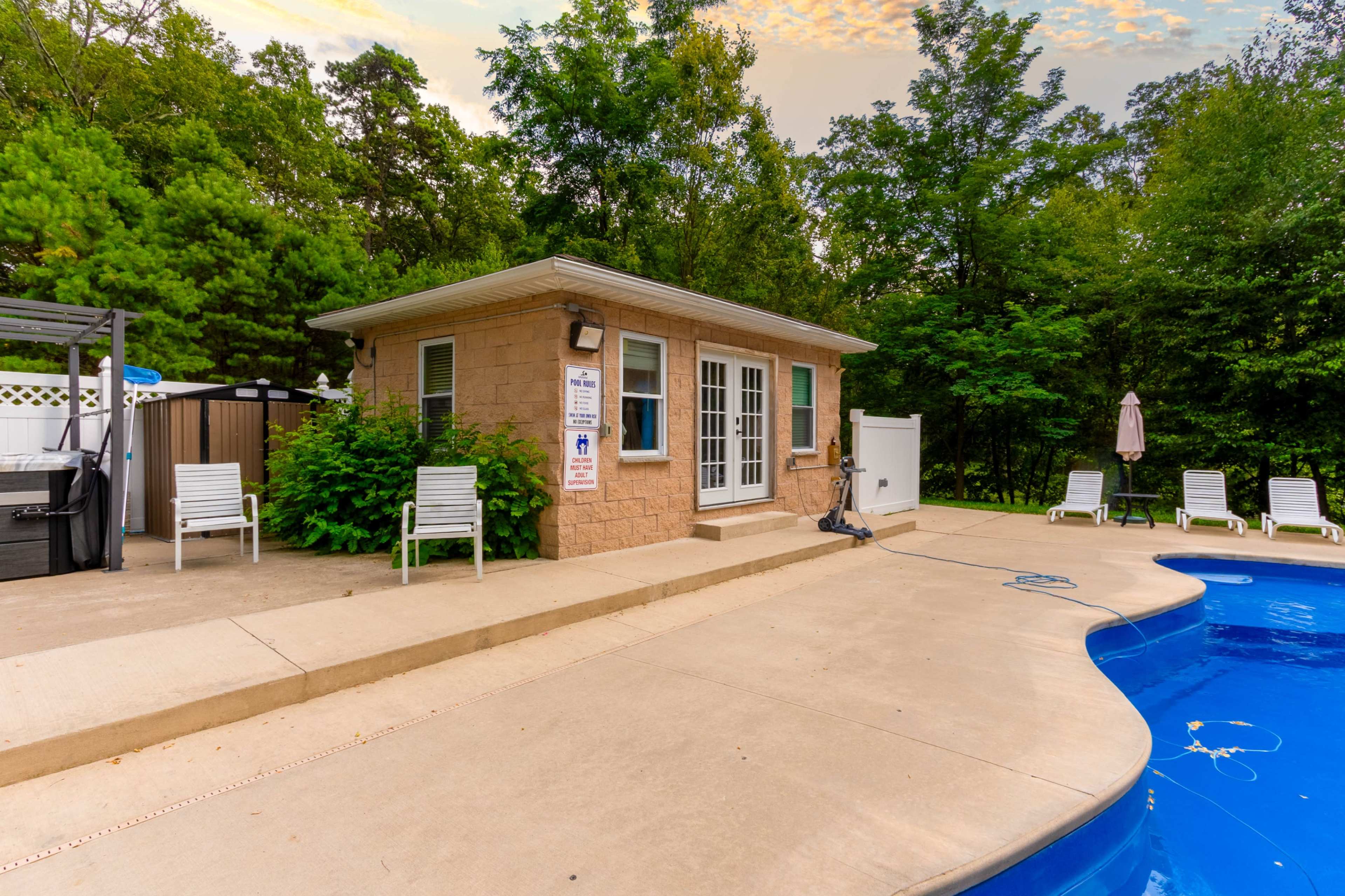 The image shows a pool area with a stone building, several lounge chairs, and a swimming pool surrounded by green trees.