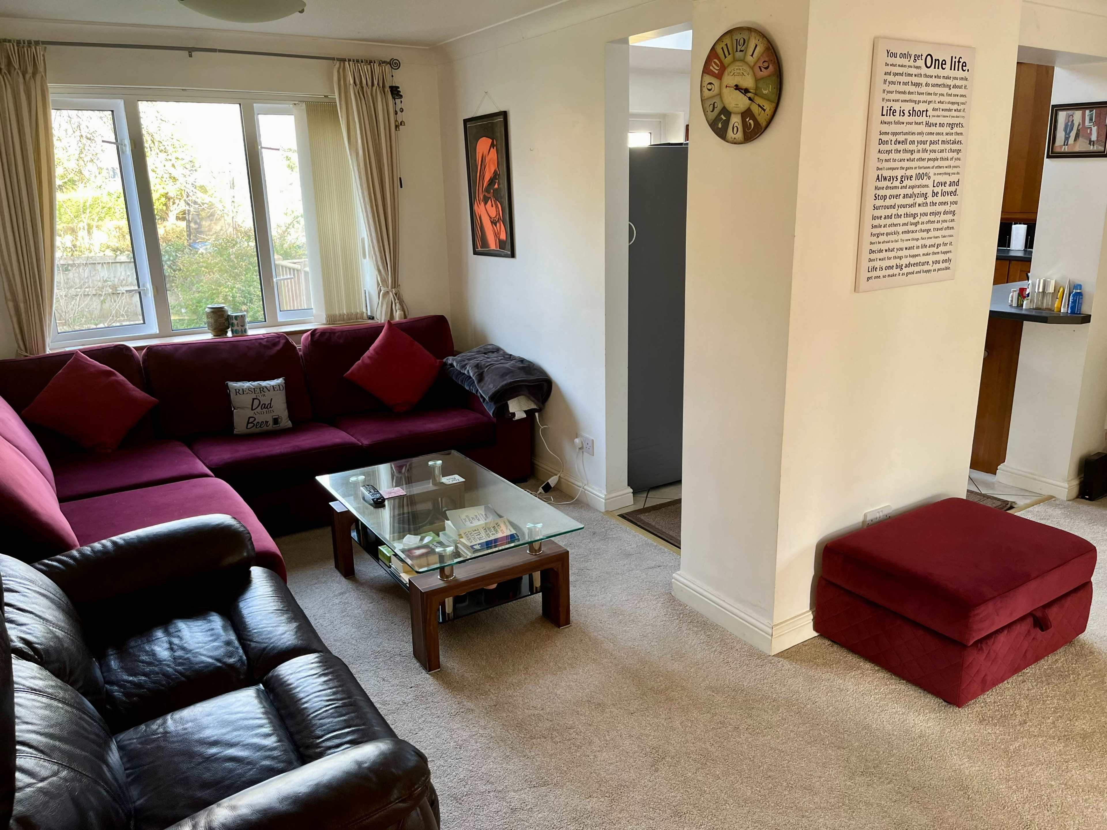 A cozy living room with a dark leather sofa, a glass coffee table, and a red cushioned ottoman, connected to an open kitchen area.