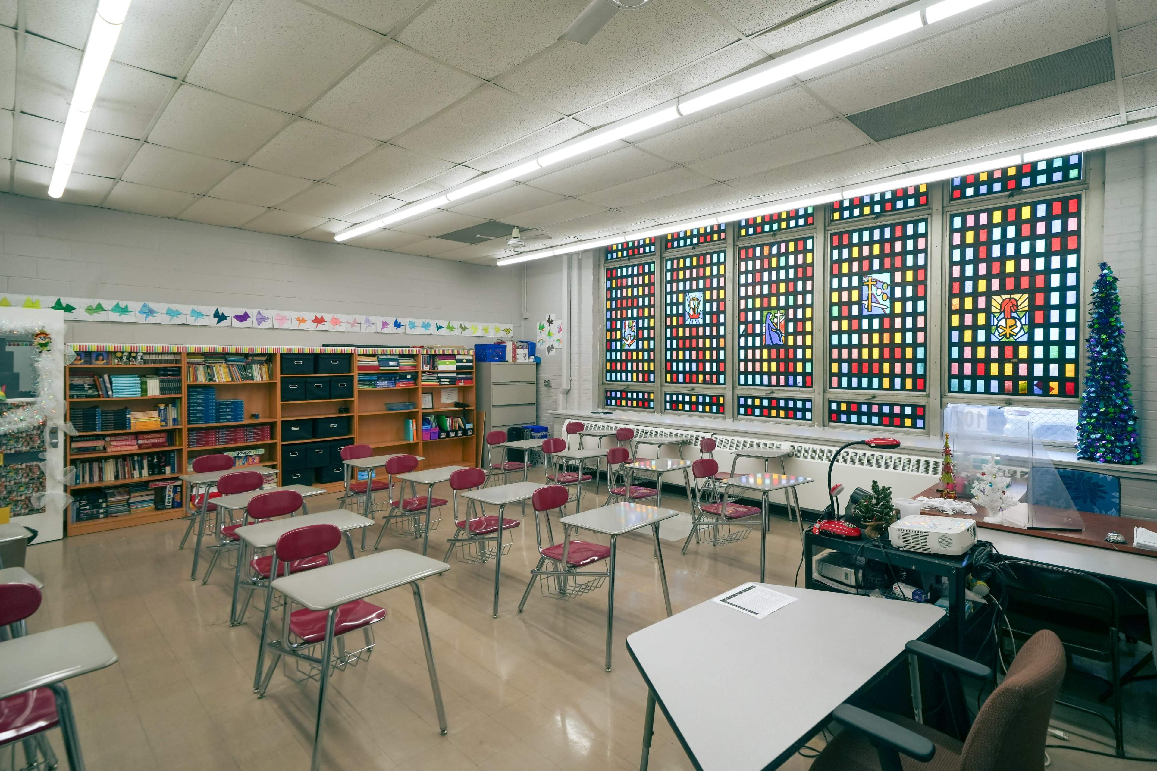 A brightly lit classroom features rows of empty desks, colorful stained glass windows, and shelves filled with books and supplies.