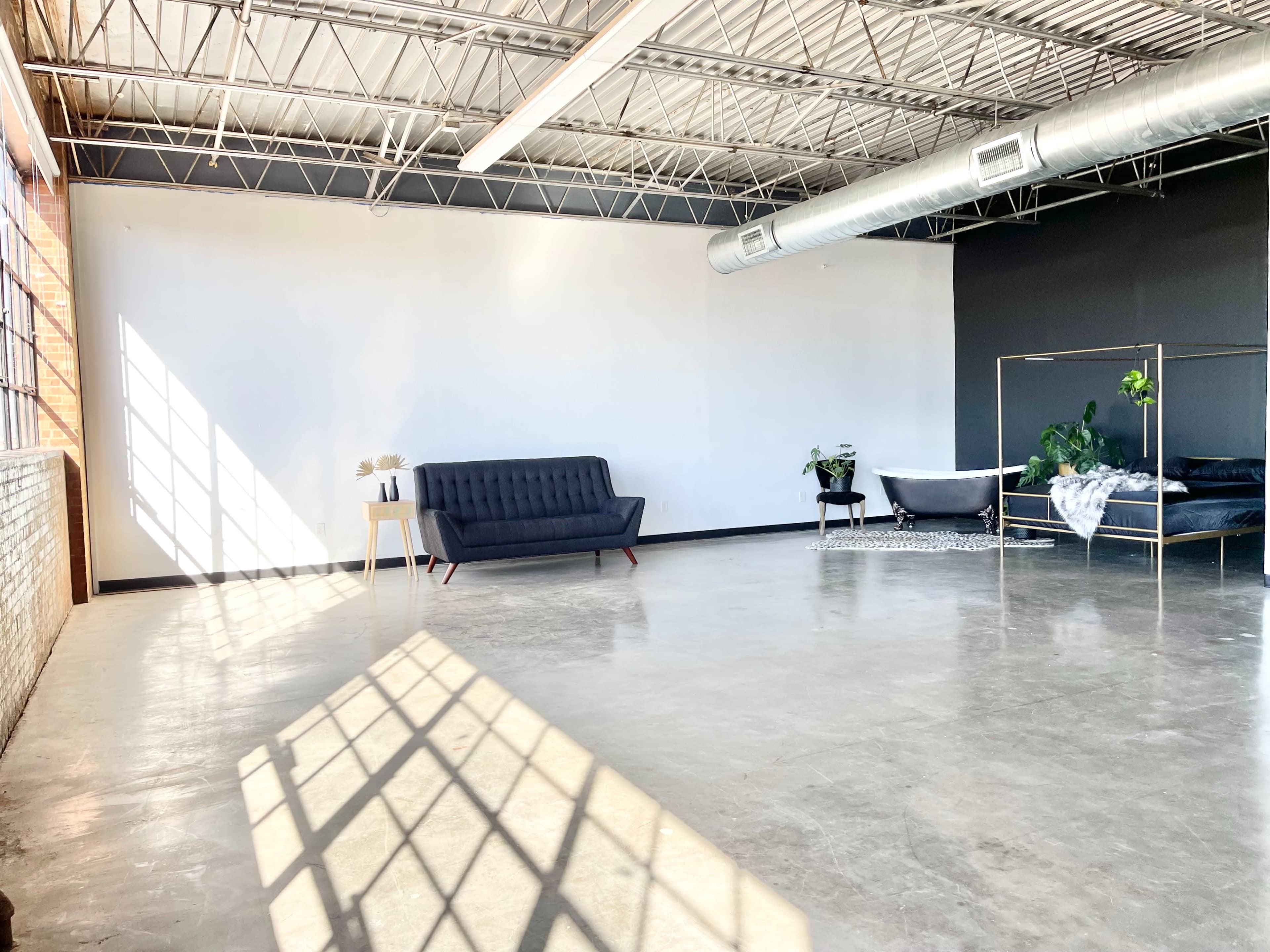 A spacious, minimalist room with large windows, a black sofa, and a bed against contrasting walls, featuring polished concrete floors and plants in the decor.