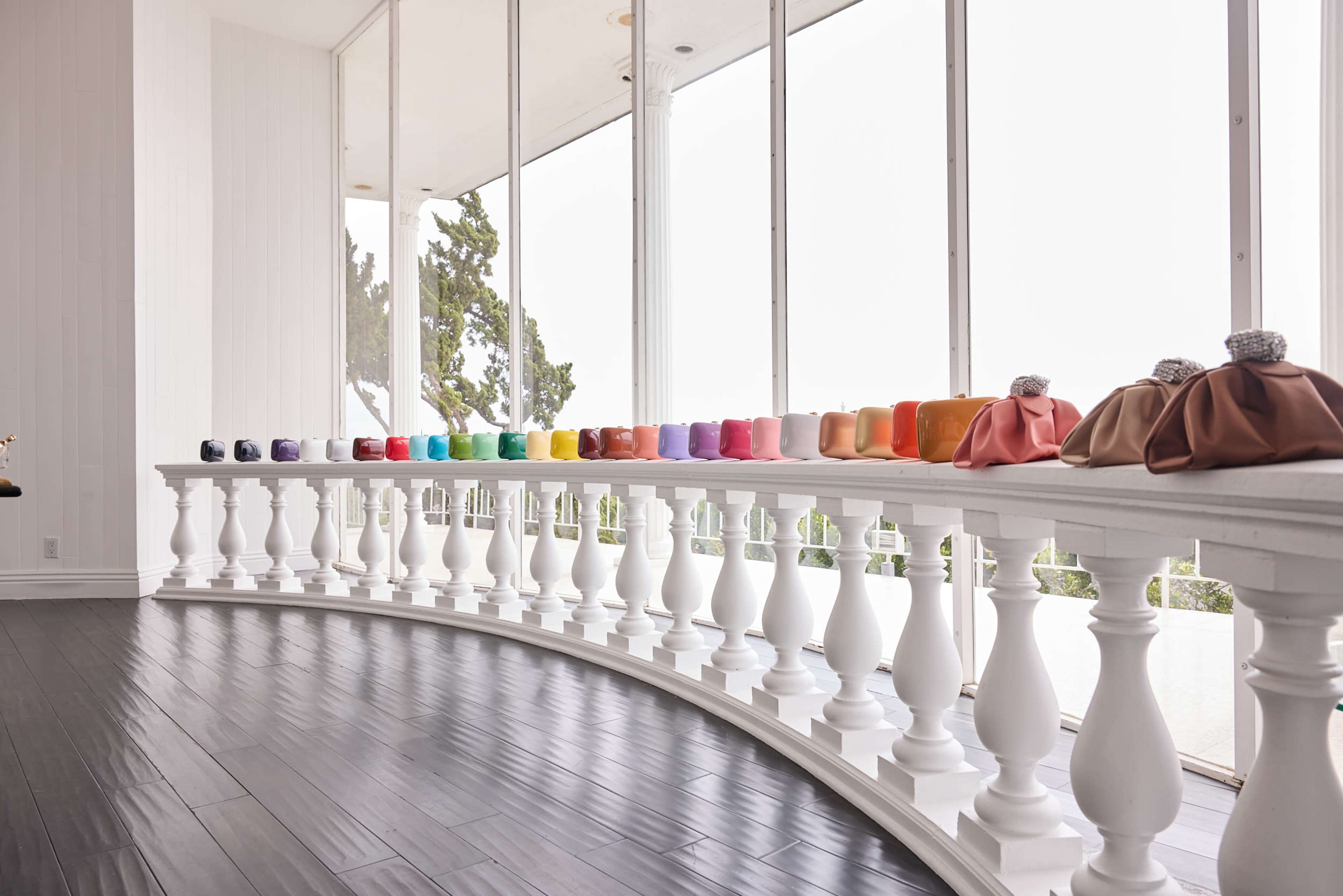 A row of colorful handbags is displayed on a white railing with large windows in the background.