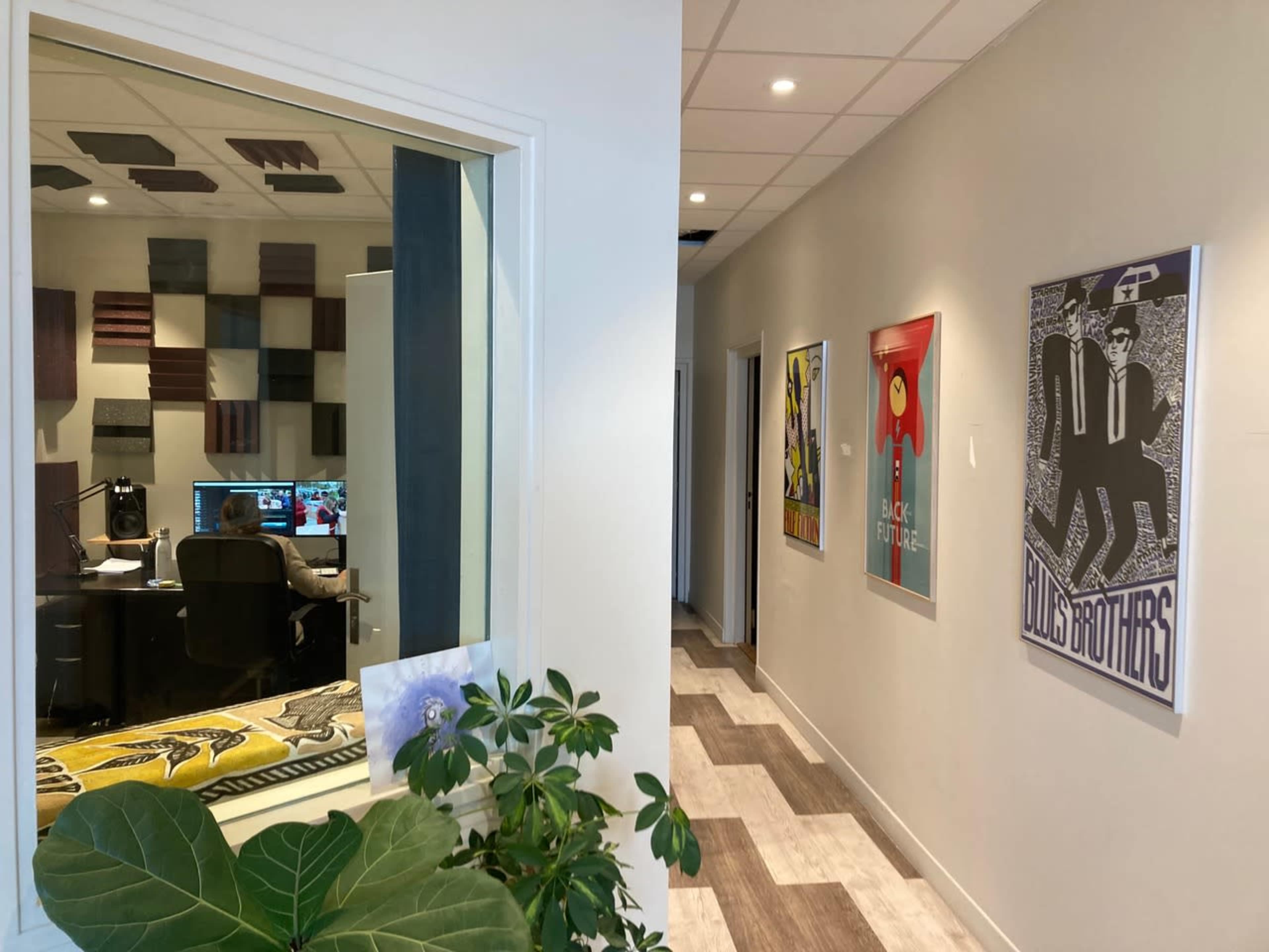 A hallway with framed posters on the walls and a window into a room with a computer setup behind it.