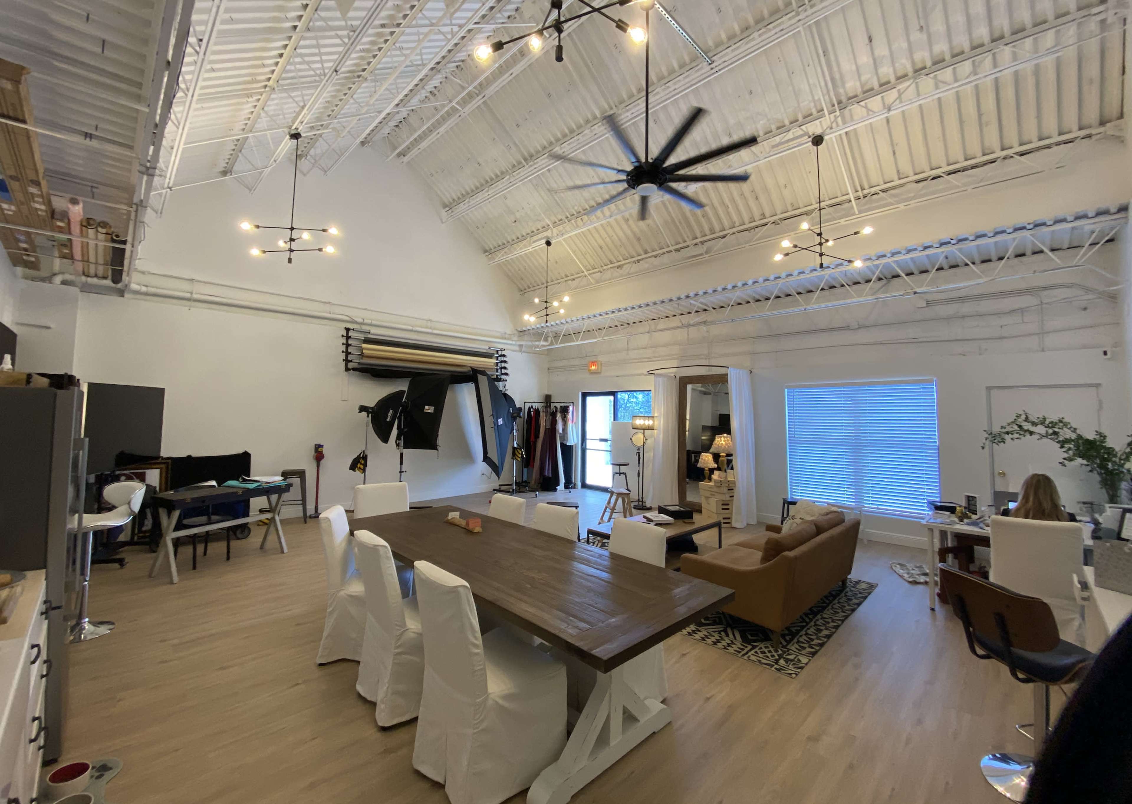 The image shows a spacious, well-lit studio with high ceilings, featuring a large wooden table, comfortable seating, and various photography equipment along the walls.