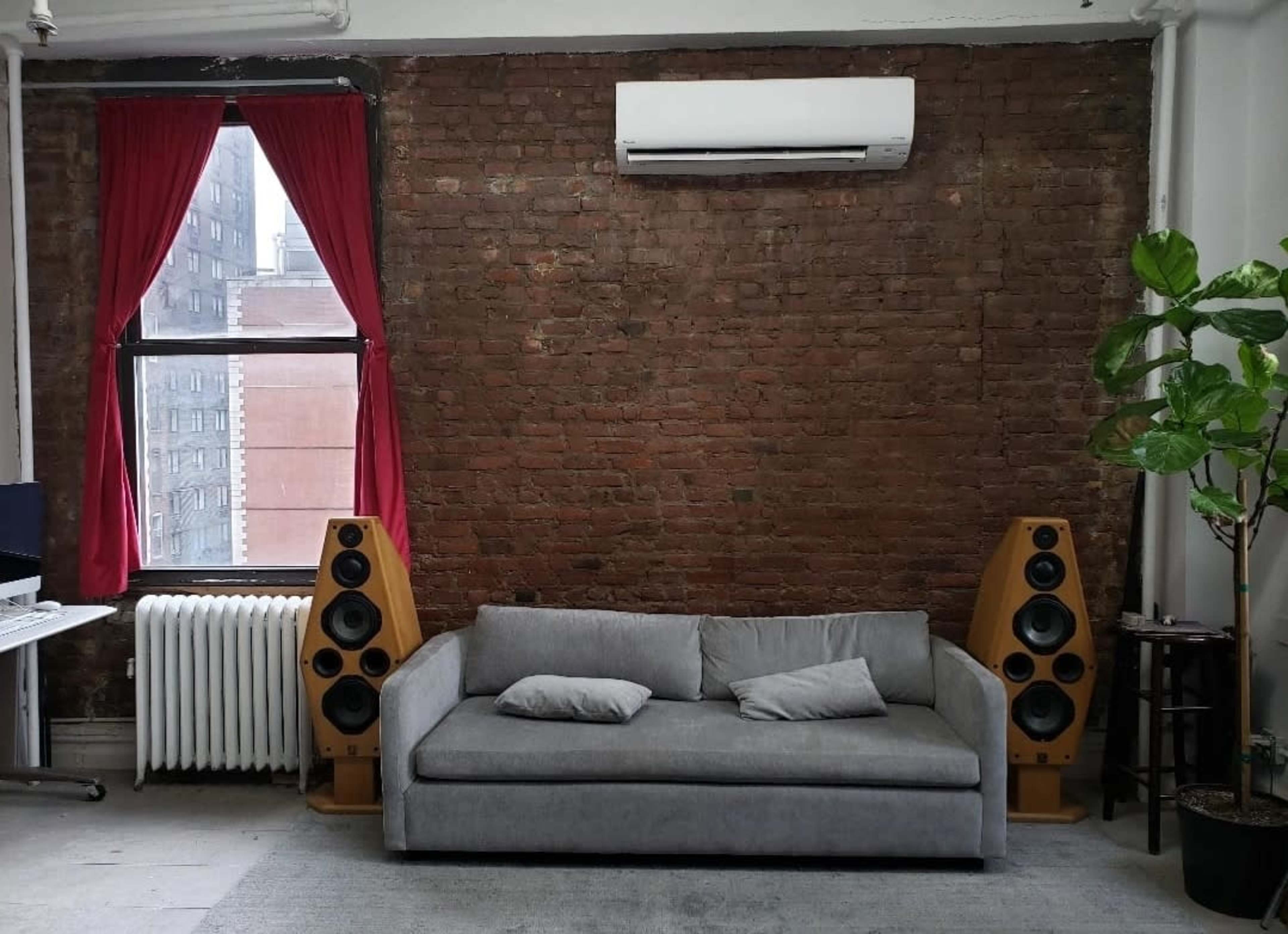 The image shows a gray sofa between two wooden speakers against a backdrop of a brick wall, with a window draped in red curtains and a potted plant to the right.