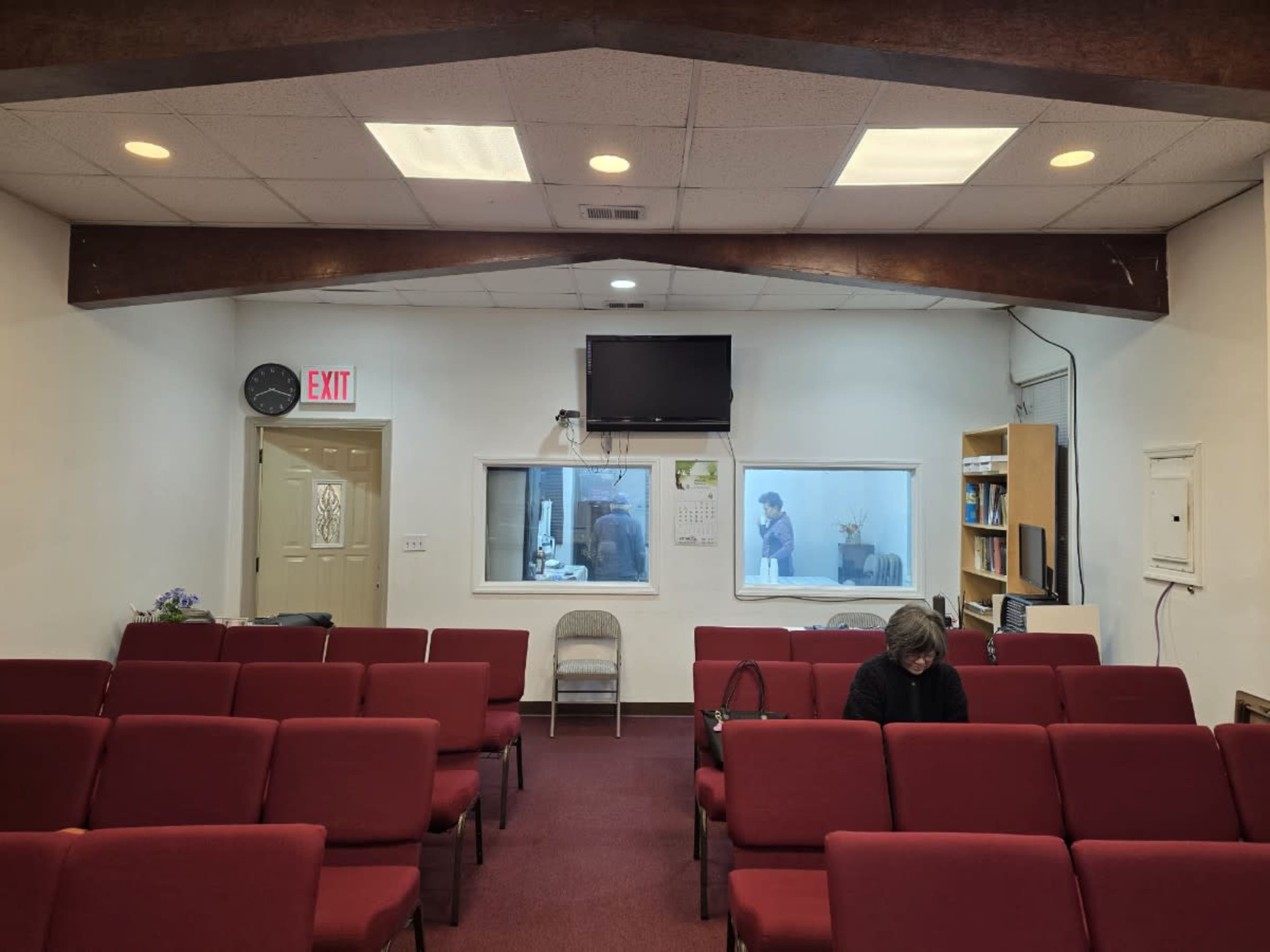 The interior of a small church or meeting room features red chairs arranged in rows, with a woman seated in front and a windowed area in the background.