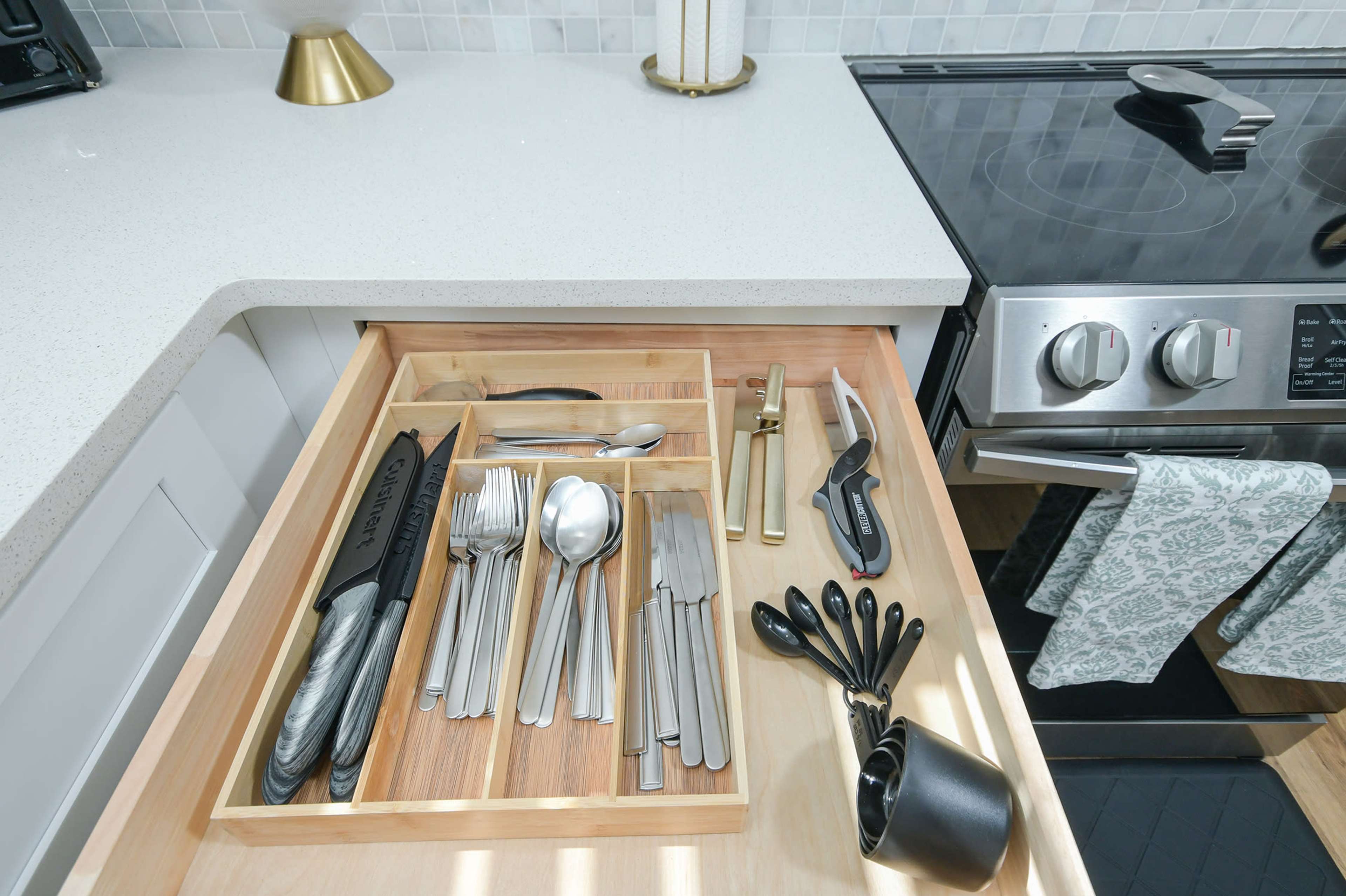 A kitchen drawer contains organized utensils, including knives, forks, spoons, and measuring spoons, arranged in a wooden tray.