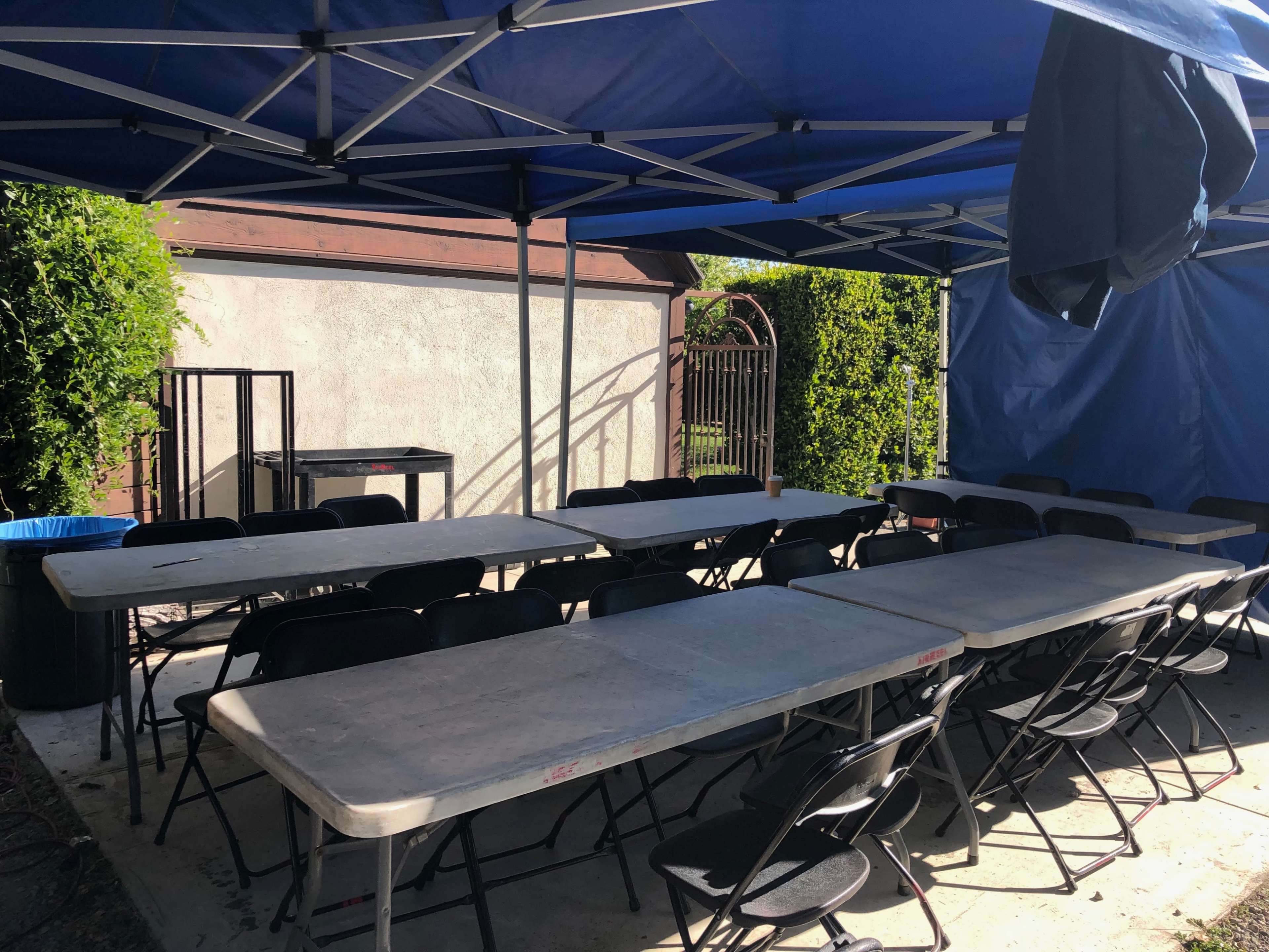 A covered outdoor area features several rows of tables and chairs arranged on a concrete surface, with a gate and greenery in the background.