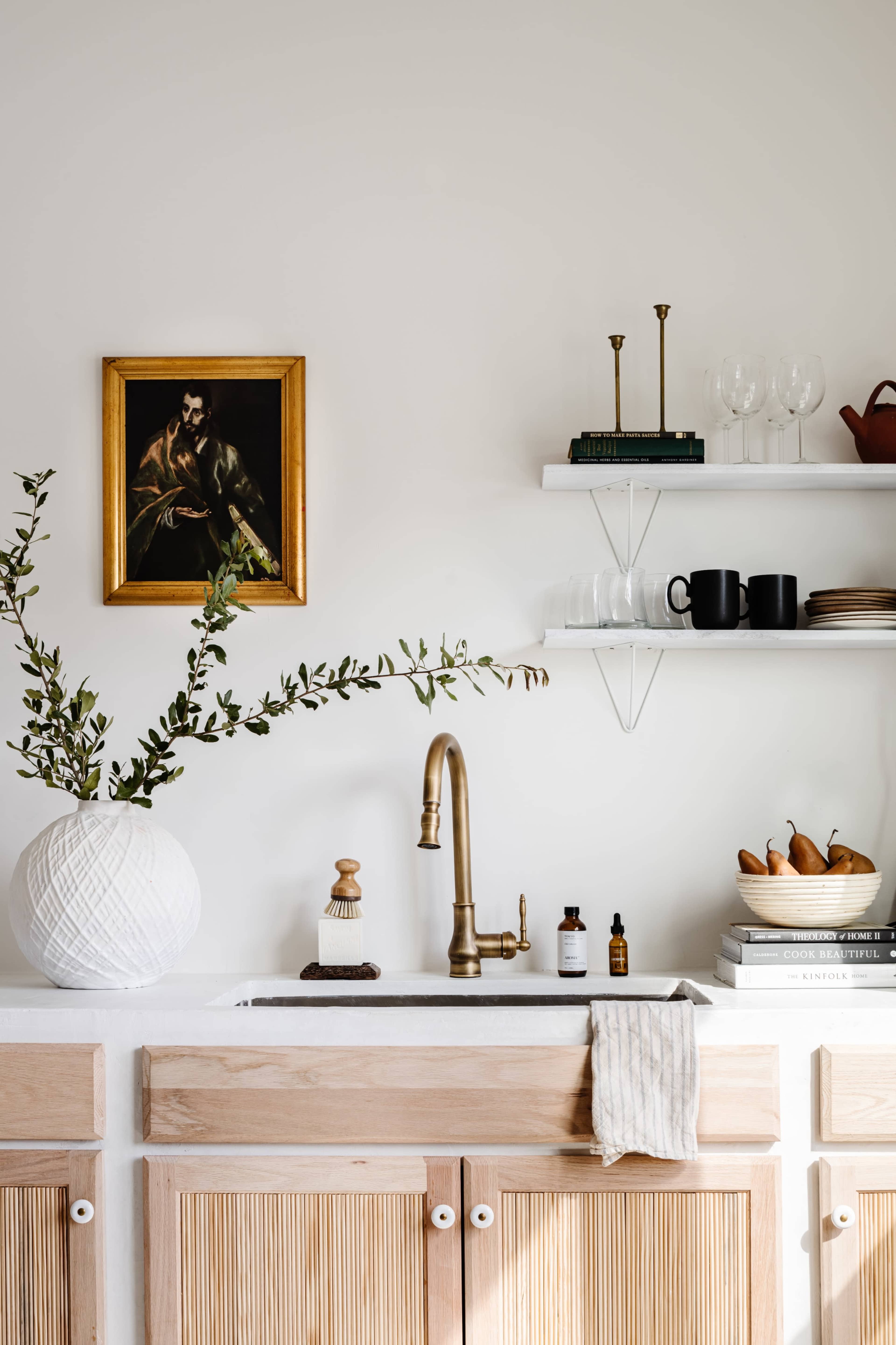 A modern kitchen features a white sink with a brass faucet, wooden cabinetry, and shelves displaying glassware, fruit, and decorative items, along with a framed painting on the wall.