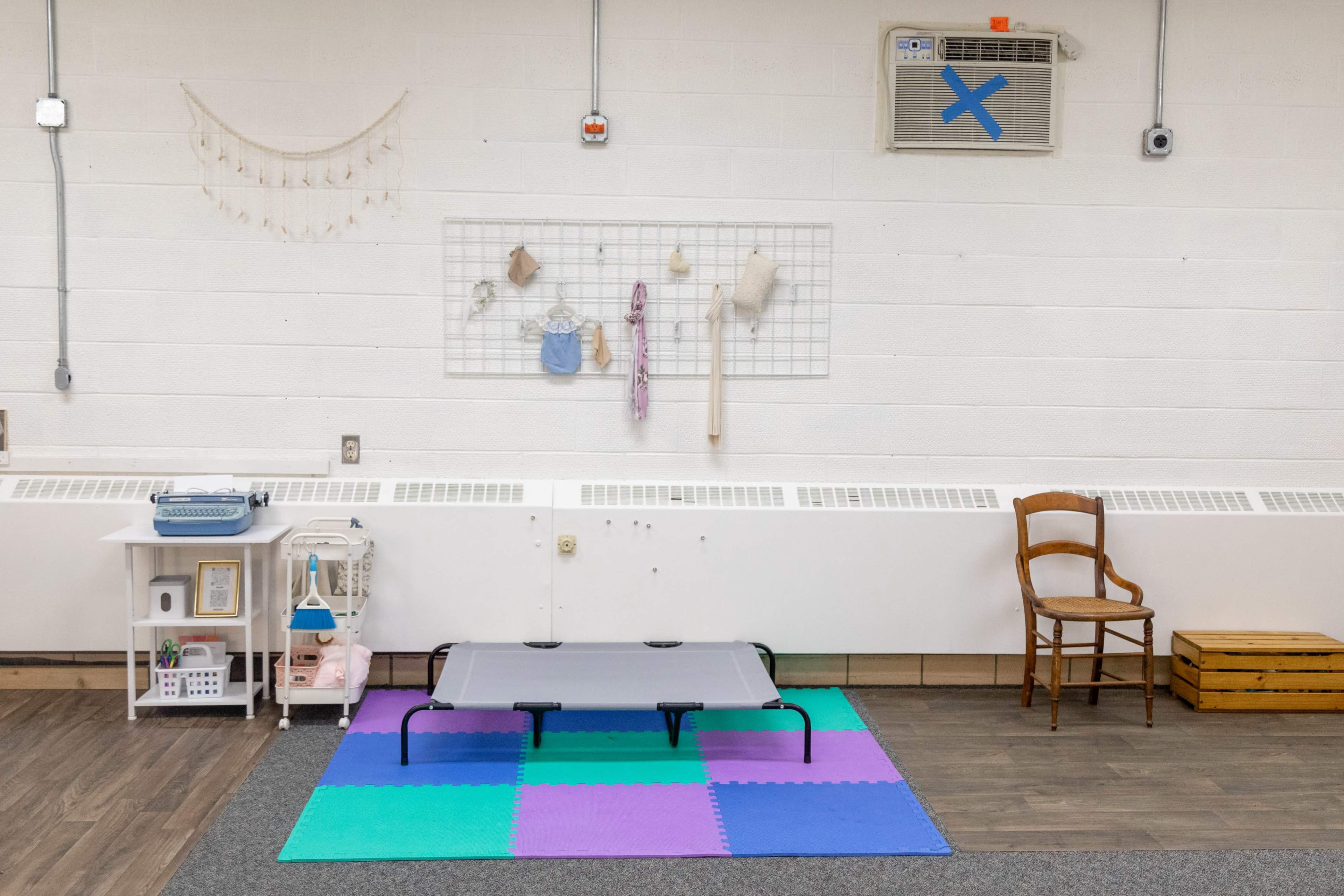 A small room features a colorful foam mat, a raised cot, a chair, and various items hanging on a wall grid.