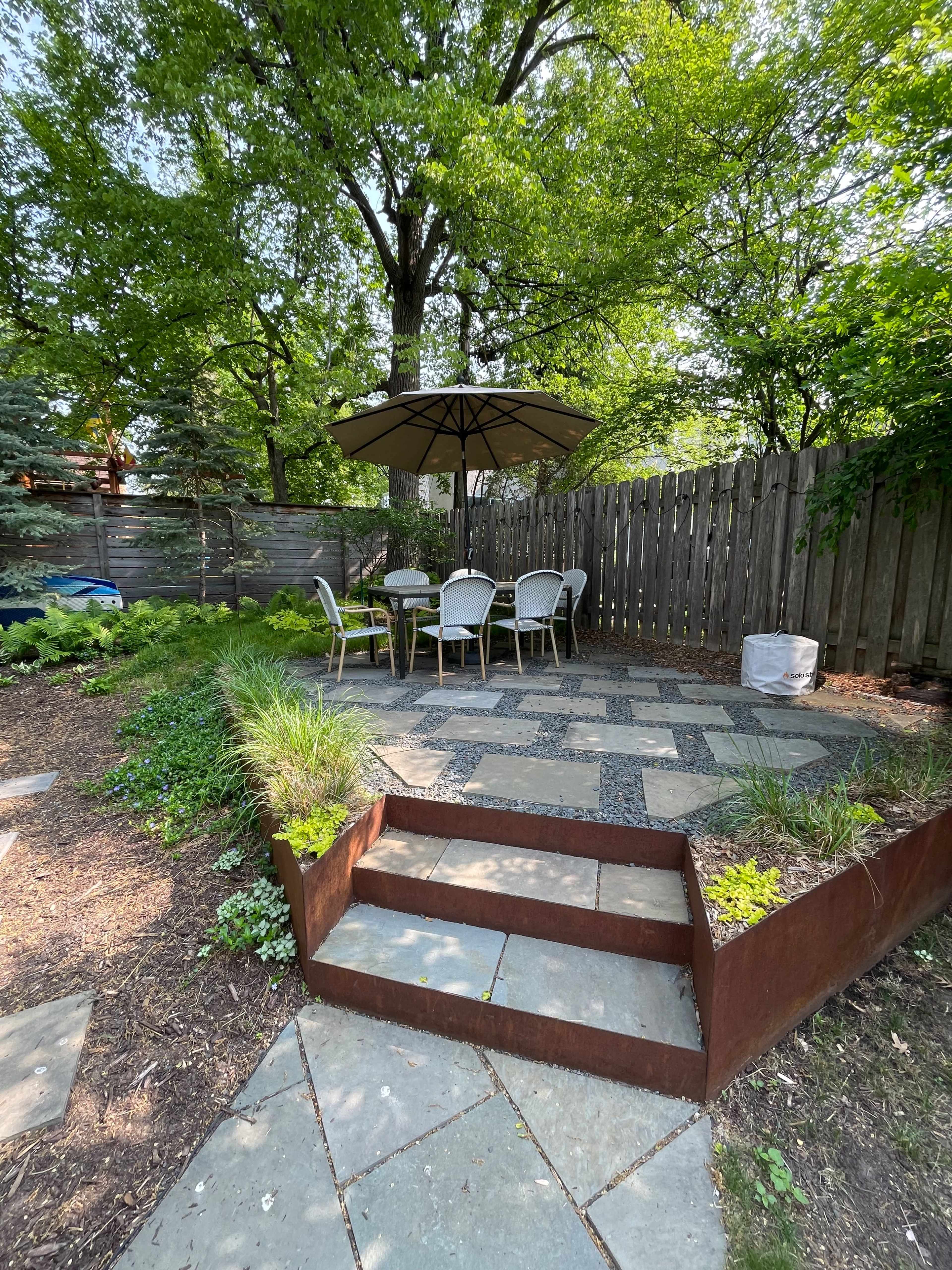 A patio area with a table and chairs is shaded by an umbrella, surrounded by greenery and bordered by a wooden fence.