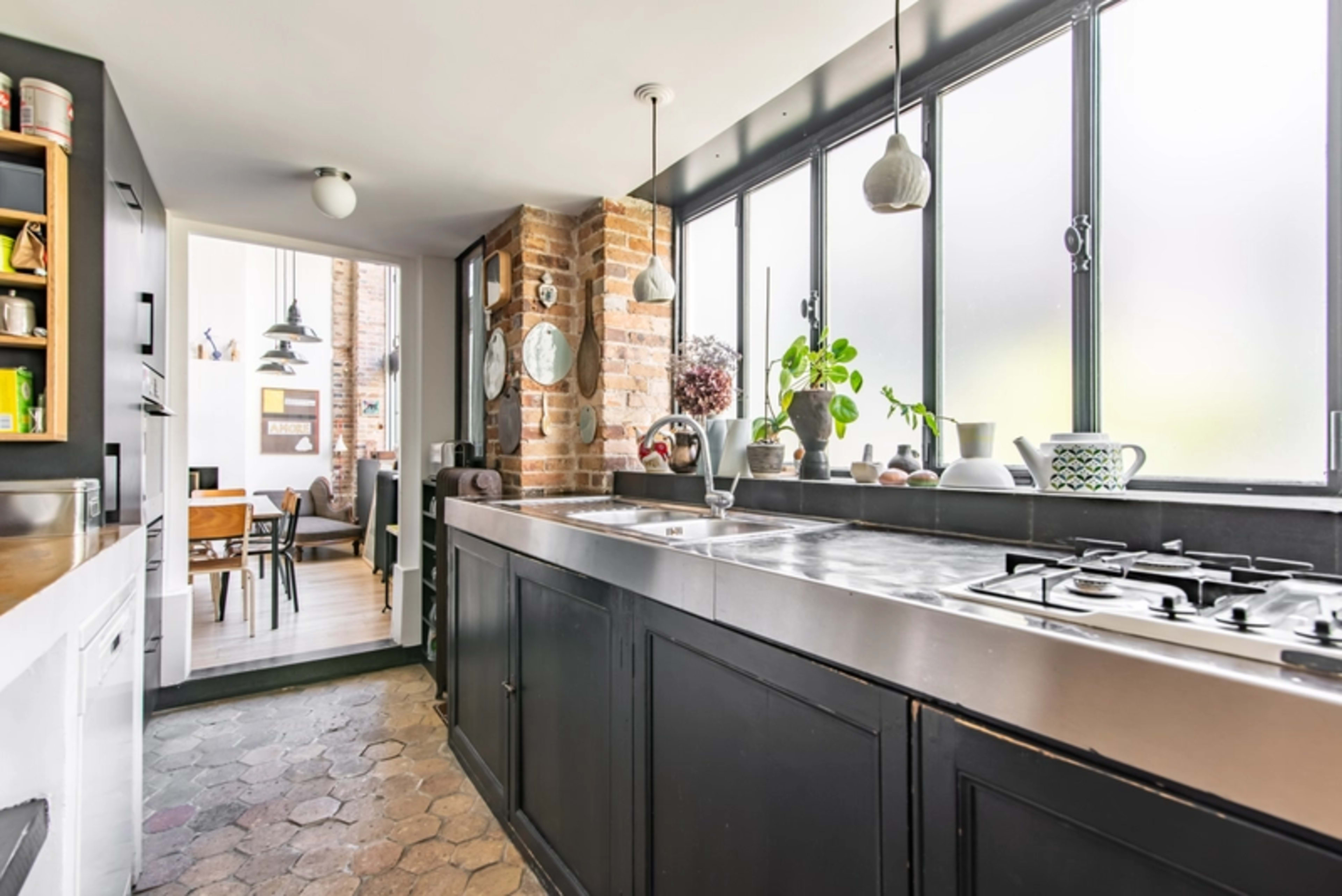A modern kitchen with a black cabinetry, a stainless steel countertop, and large windows allowing natural light to enter.