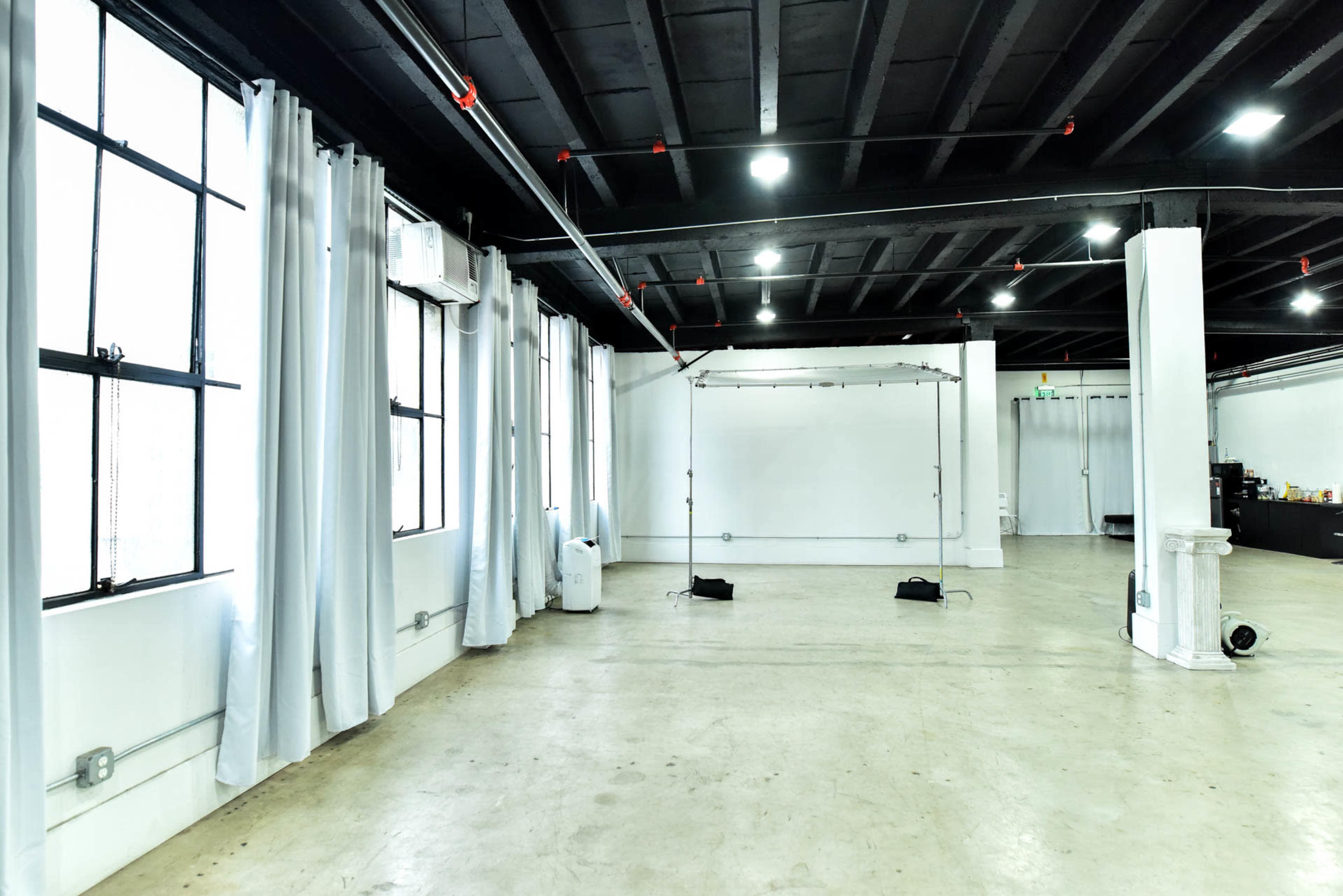 The image shows an empty, spacious studio with windows, concrete flooring, and a white wall at the far end.