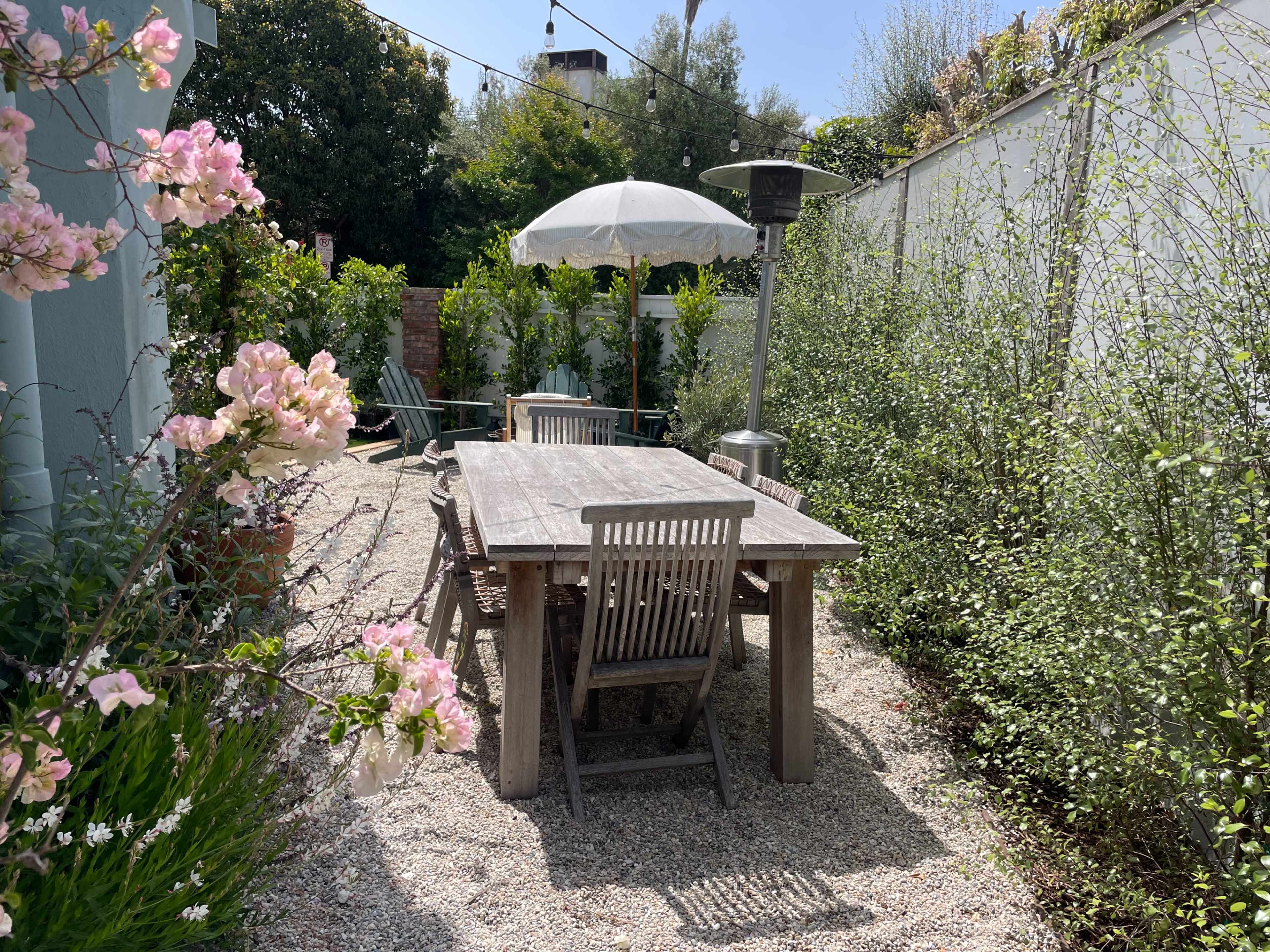 A wooden table with chairs is positioned under an umbrella in a gravel garden surrounded by plants and flowers.