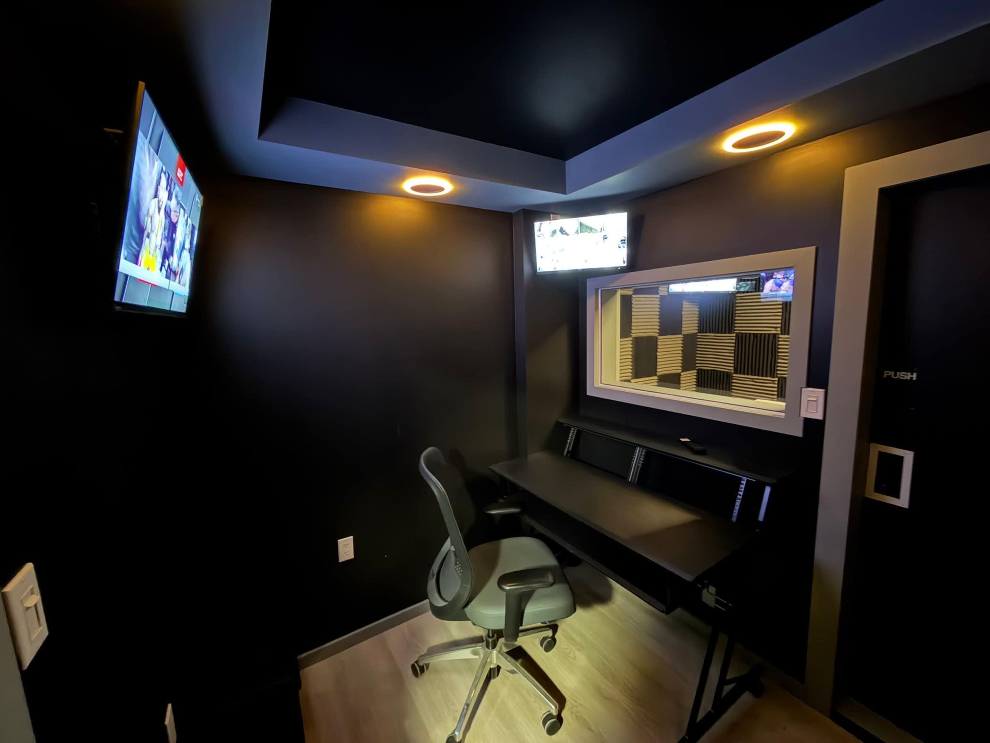 The image shows a small, modern workspace featuring a desk and rolling chair, with two wall-mounted screens displaying content and a window looking into a soundproof room.
