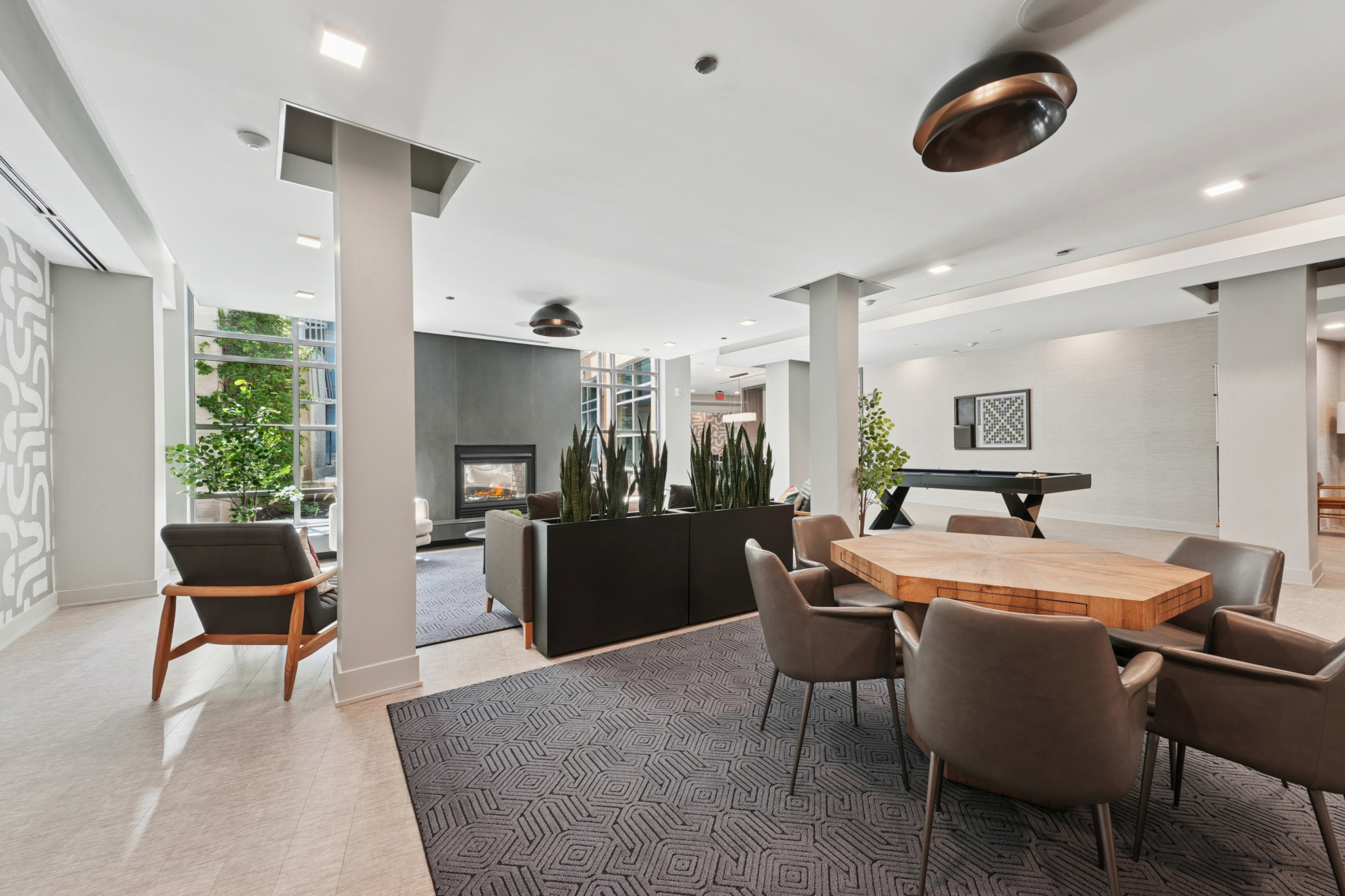 The image shows a modern communal lounge area featuring a seating arrangement with chairs and a wooden table, plants, and a fireplace.