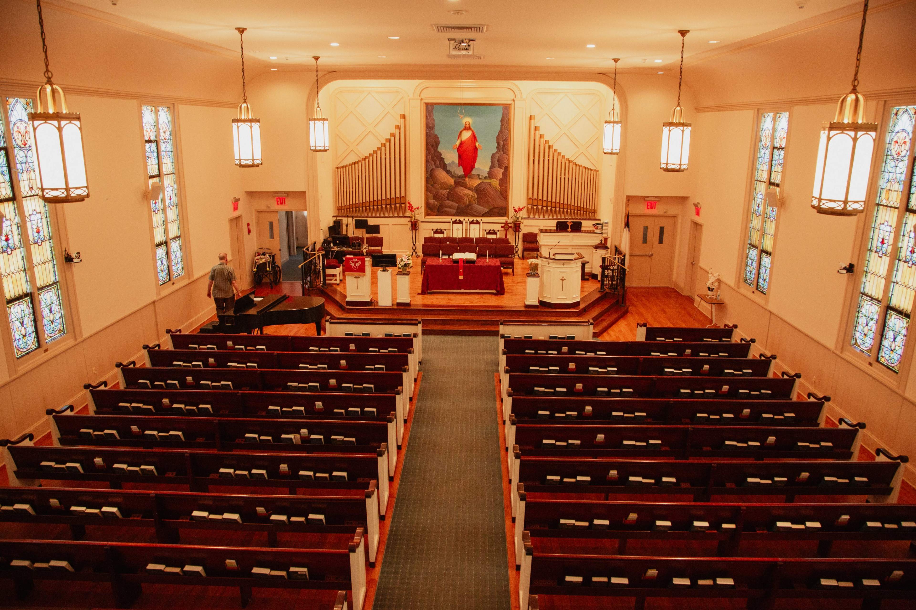 The image shows the interior of a church, featuring rows of wood pews, stained glass windows, an altar with a large painting, and a pipe organ.