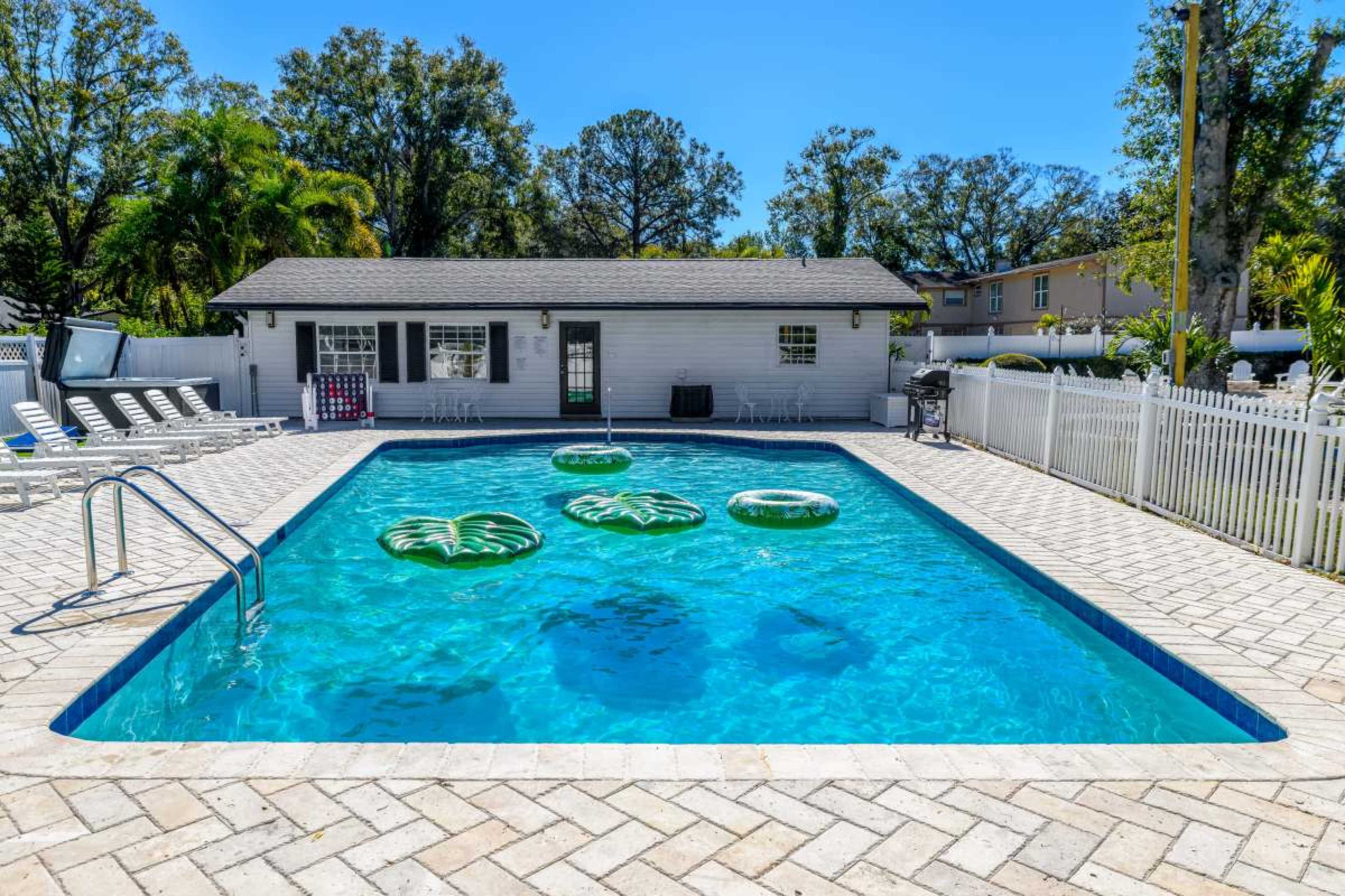 The image shows a rectangular swimming pool with green inflatable floaties and a fenced area containing lounge chairs and a building in the background.