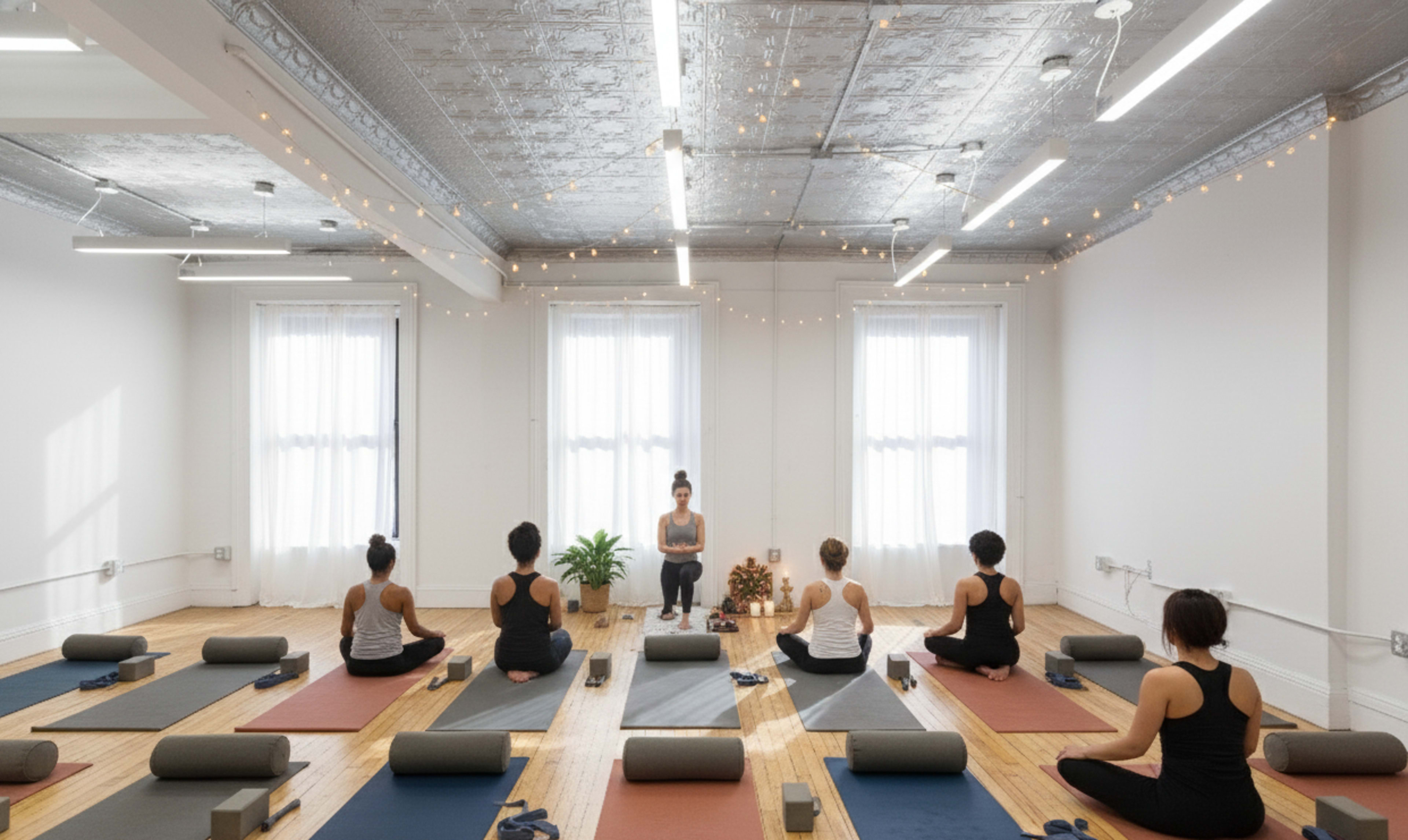 A yoga studio features several practitioners seated on mats, facing an instructor at the front, with string lights illuminating the space.