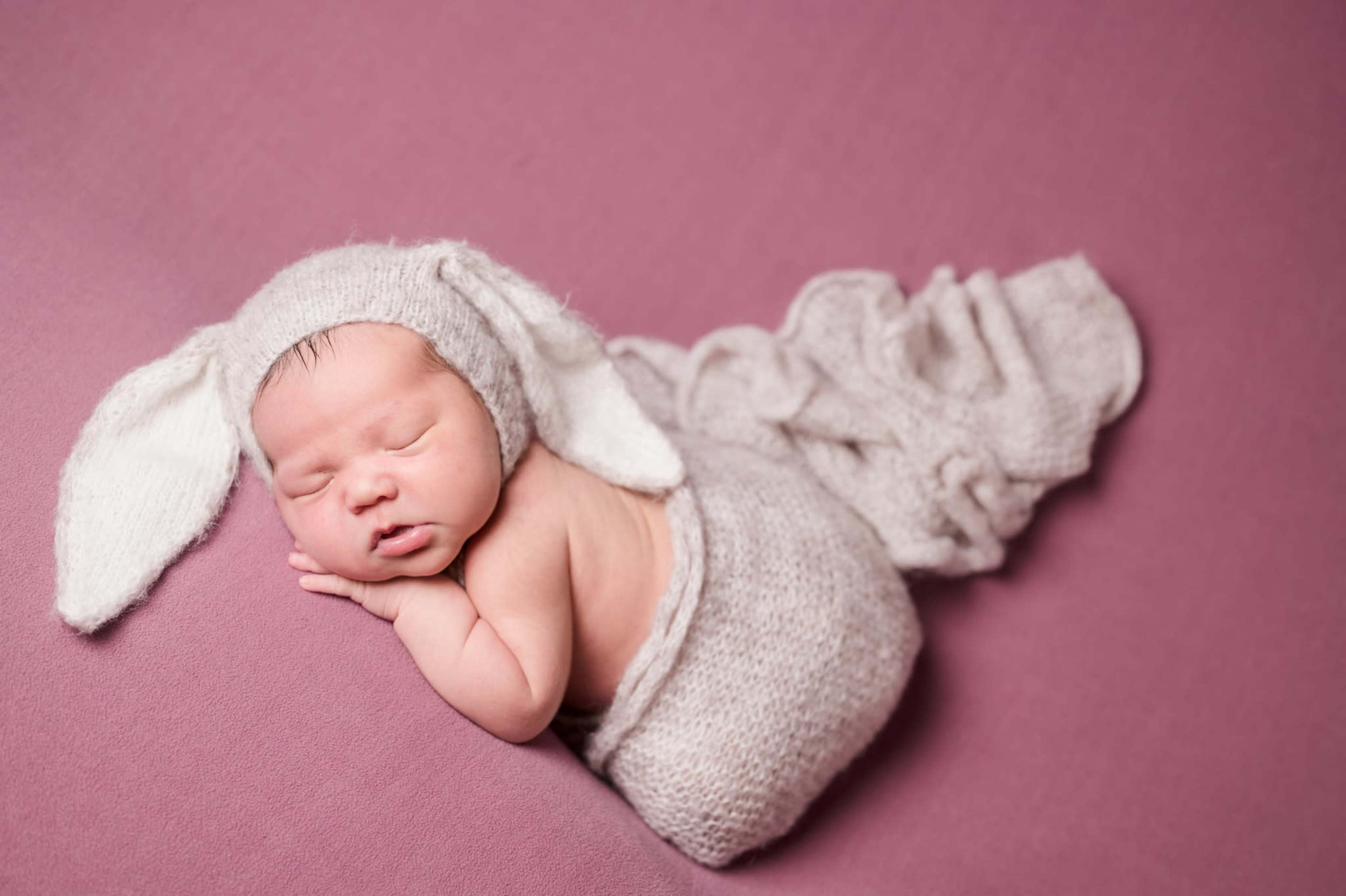 A newborn baby is peacefully sleeping on a pink background while wrapped in a knitted fabric with bunny ears.