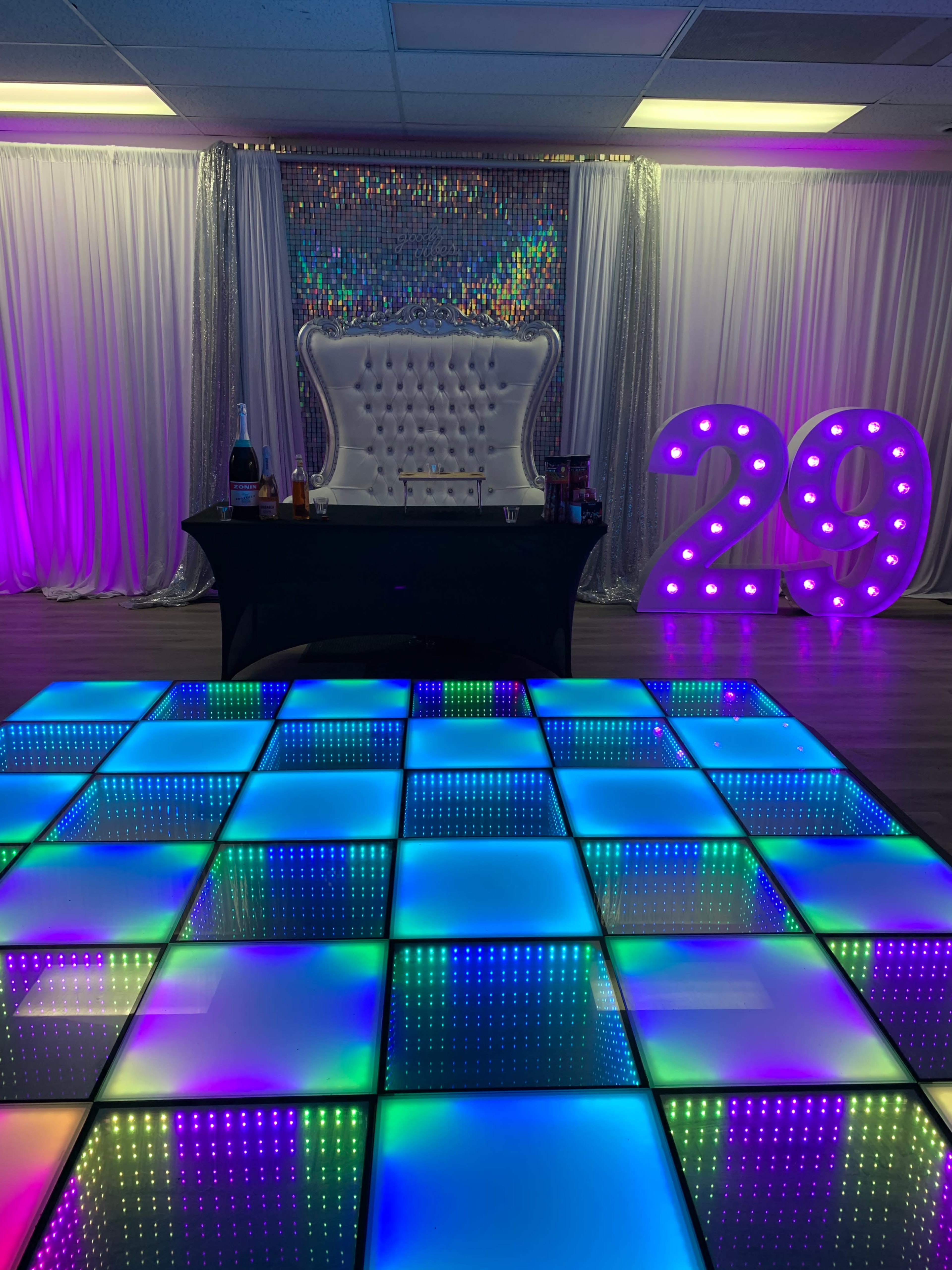 A decorated party space featuring a light-up dance floor, a bar setup, and a large illuminated "29" display against a backdrop of shimmering curtains.